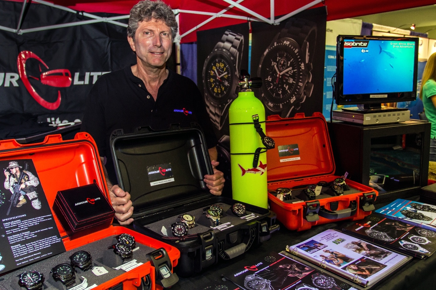 Ashley Diener, founder and owner of ArmourLite Watches, stands behind a trade show booth displaying an array of ArmourLite watches on the counter. He is speaking to attendees and sharing information about the brand’s durable, tactical, and illuminated timepieces. The booth is professionally arranged with watches in cases, promotional materials, and branding visible. This image introduces the article on Ashley Diener’s interview with The Armory Life, providing context for the discussion on watchmaking, innovation, and the ArmourLite brand. Trade show visitors are visible in the background, emphasizing the event setting. The photo sets a professional and approachable tone for the article and connects readers to the subject matter.