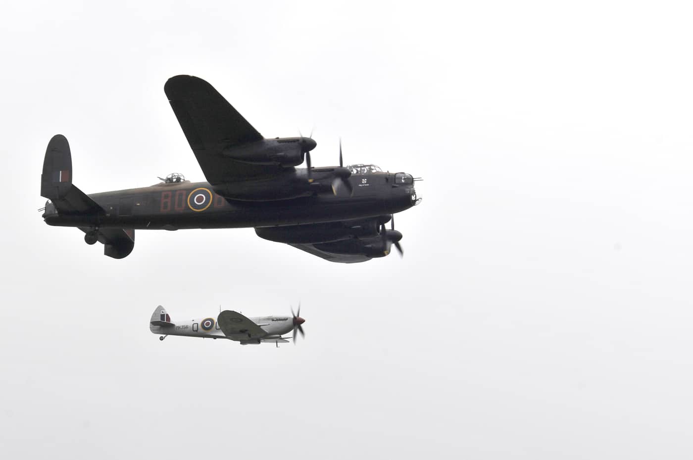 This photo captures an Avro Lancaster heavy bomber flying in formation with a Supermarine Spitfire single-seat fighter during the Battle of Britain Memorial Flight in 2012. The aircraft are shown mid-air, both painted in authentic RAF wartime colors and markings, including roundels and squadron codes. The Lancaster, much larger with four engines and a twin tail, is flying alongside the more compact and agile Spitfire. The sky is bright and clear, ideal for an air display, and both planes are well lit, allowing details such as the Lancaster’s turrets and the Spitfire’s elliptical wings to be clearly seen. The Battle of Britain Memorial Flight is an annual event that features flying displays with restored WWII aircraft, serving as a tribute to RAF aircrew and their contributions. The image emphasizes the contrast in size and design between the two planes. The photo highlights both technical heritage and the ongoing public commemoration of Britain’s aerial history.