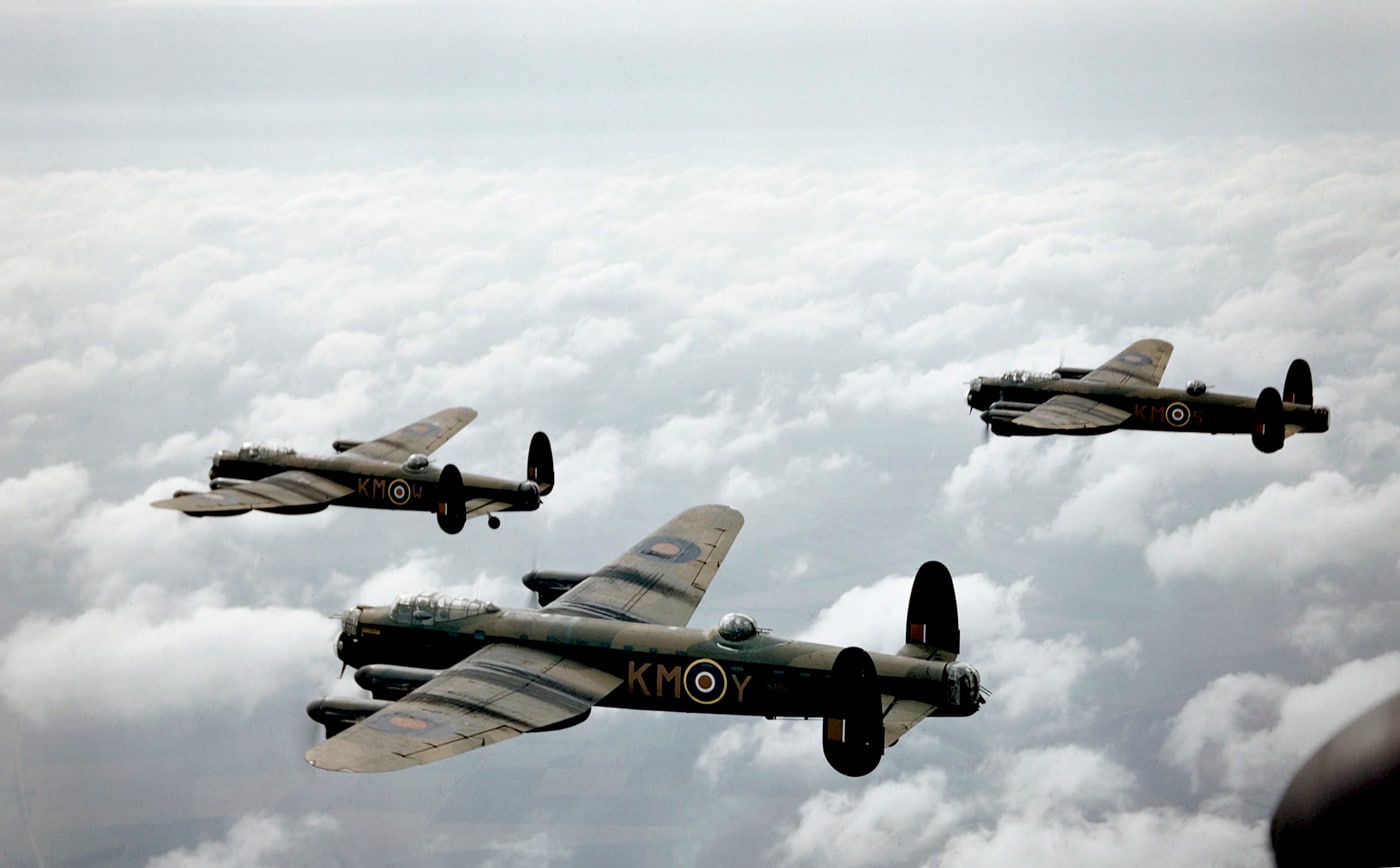 The image is a color photograph taken in September 1942, showing several Avro Lancaster bombers in flight above the British countryside. The Lancasters are flying in formation, each displaying wartime camouflage paint and RAF roundels typical of the period. The color photo highlights details such as the green and brown upper surfaces and the pale undersides, as well as markings like squadron codes and aircraft serial numbers. The bombers’ four engines and large wingspans are clearly visible, with sunlight reflecting off the metal surfaces and glass cockpits. The background includes blue sky and scattered clouds, providing a sharp contrast to the aircraft. This rare color image gives an authentic representation of how the Lancasters appeared in operational service during the early years of WWII. The photo is historically valuable for illustrating articles about wartime aviation, Bomber Command, and the Avro Lancaster’s active missions in 1942.