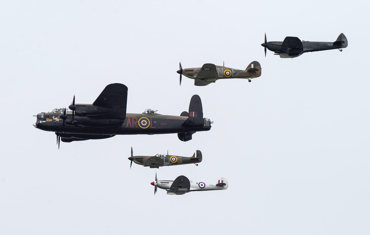 The image shows an Avro Lancaster heavy bomber in flight, escorted by four Supermarine Spitfire fighters, all flying in a close and organized formation during an air show. The Lancaster, with its four engines and distinctive silhouette, takes center position, while the Spitfires — smaller, single-seat fighters — fly evenly spaced, two on each side. The sky is clear or partly cloudy, providing good visibility of all aircraft. Spectators on the ground may be faintly visible, looking up as the formation passes overhead. The photograph highlights the enduring legacy and public admiration of Britain’s WWII aircraft, with the Lancaster and Spitfires representing the Royal Air Force’s power and teamwork. No combat context is present; the scene is peaceful, commemorative, and celebratory. This image captures the essence of air shows that honor RAF history and the iconic roles of the Lancaster and Spitfire.
