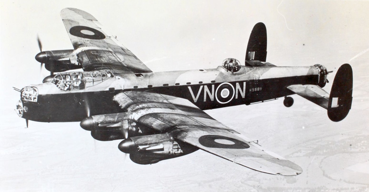 A lead image shows an Avro Lancaster heavy bomber in flight, photographed from the side against a backdrop of blue sky. The plane’s four Rolls-Royce Merlin engines and distinctive twin tailfins are clearly visible. The aircraft’s long, straight wings are prominent, underscoring its reputation for carrying large bomb loads during World War II. This general photo can represent any stage of the Lancaster’s career, from early missions to its later use in famous operations like the Dambusters raid. The photo’s perspective allows for easy identification of the Lancaster’s main features, such as its mid-upper turret and broad fuselage. The overall composition is simple and timeless, making it a suitable lead image for any discussion of the Avro Lancaster. The image introduces the article and orients the reader to the topic of RAF heavy bombers.