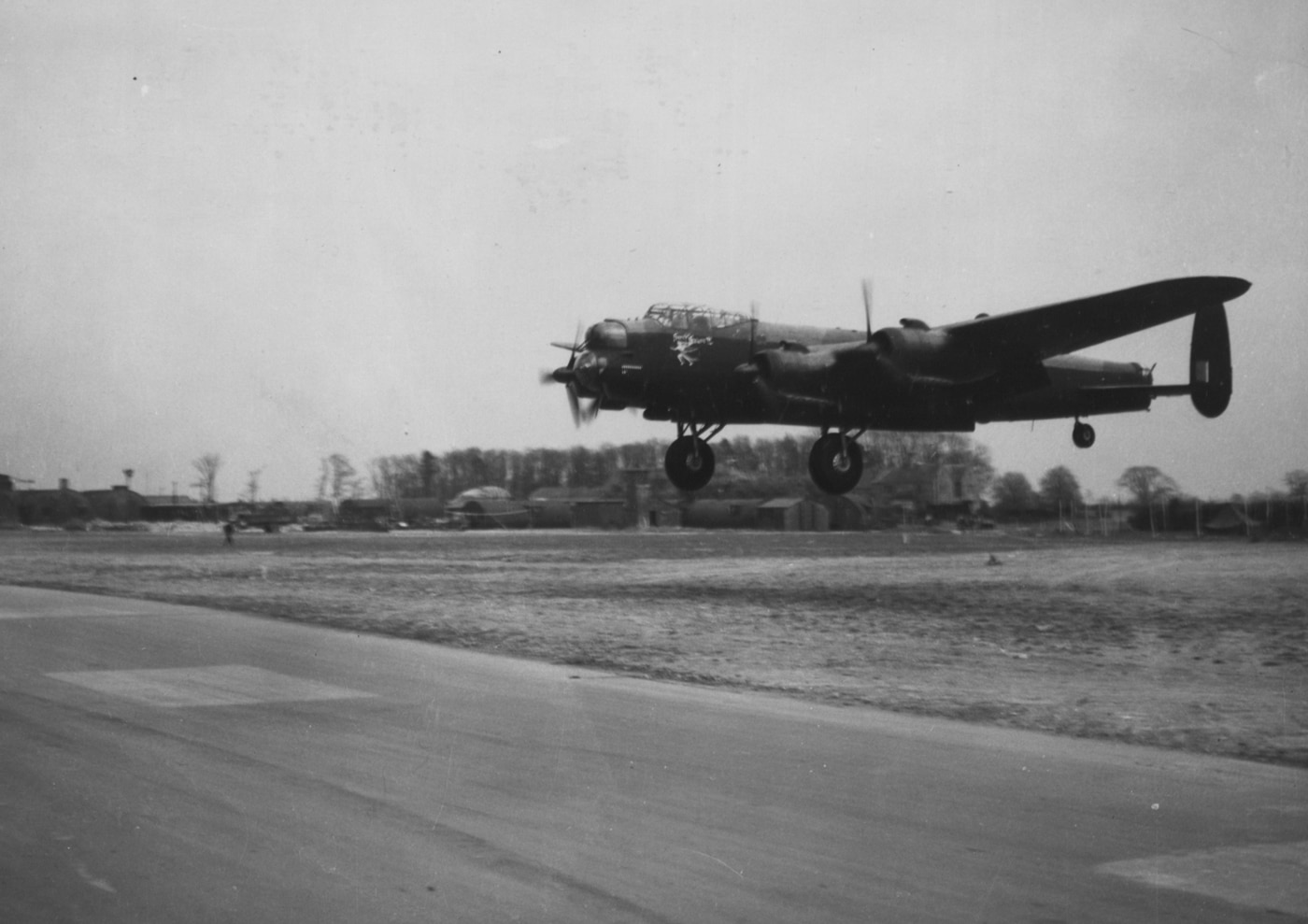 The photograph shows an Avro Lancaster heavy bomber in the process of landing on a runway at a US 8th Air Force base, most likely located in England during World War II. The Lancaster’s main wheels are just touching down or rolling along the tarmac, with the aircraft’s nose slightly raised as it completes its approach. The background includes American-style hangars, ground vehicles, or support equipment typical of a USAAF base. The Lancaster’s four engines, distinctive twin tailfins, and RAF markings are clearly visible, indicating the British origin of the aircraft. This landing scene illustrates the close operational ties between the Royal Air Force and the United States Army Air Forces, who frequently shared airfields and collaborated on strategic bombing missions. The lighting and setting provide a sense of wartime activity and Allied cooperation. The photo is suitable for illustrating articles about Allied air strategy, base-sharing, or the Lancaster’s international role.