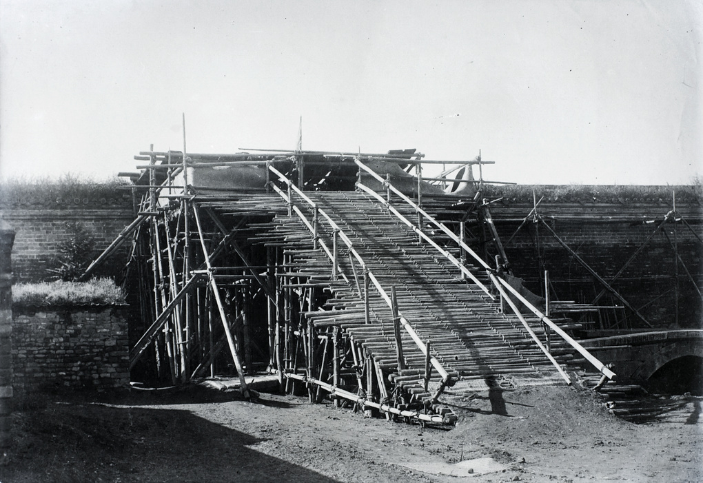 An elevated wooden platform or siege tower, hastily built by Boxer rebels, stands just a few feet from the massive stone Tartar Wall in Peking during the Boxer Rebellion. The structure is constructed of bamboo poles, wooden planks, and rope, rising high enough for attackers to shoot directly into the defensive positions held by U.S. Marines and other allied troops below. Several Boxer fighters may be visible on the platform, armed with rifles or makeshift weapons, aiming or firing at the wall’s defenders. The background shows the imposing masonry of the Tartar Wall, with possible signs of damage or smoke from ongoing attacks. This improvised siege engine illustrates the Boxers’ resourcefulness and the intense close-quarters combat faced by Marines during the 55-day siege. The image captures the deadly standoff and the constant pressure placed on foreign defenders by innovative Boxer tactics in the crowded city.