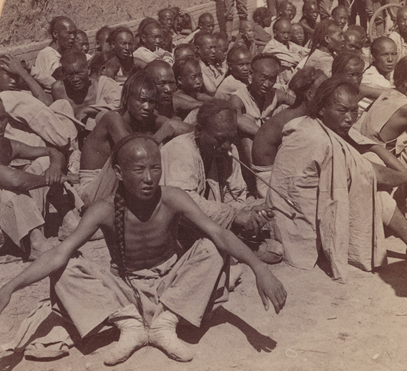 The photograph shows several Chinese Boxer prisoners seated or crouched on the ground, surrounded by armed members of the 6th U.S. Cavalry in China during the Boxer Rebellion. The prisoners are dressed in simple, worn clothing, some with hands bound or tied behind their backs. American cavalrymen in field uniforms, armed with carbines or pistols and wearing campaign hats, stand watch over the group, maintaining a tight perimeter. The setting is outdoors — likely a temporary camp or open area near a recent battle — with scattered equipment, horses, and possibly other soldiers in the background. The image documents the capture and detention of suspected Boxer fighters by U.S. forces, illustrating the process of counterinsurgency and occupation. The expressions and body language of both prisoners and guards reveal tension, exhaustion, and uncertainty, characteristic of such moments during military campaigns. This photograph captures a direct encounter between American troops and Boxer rebels in 1900 China.