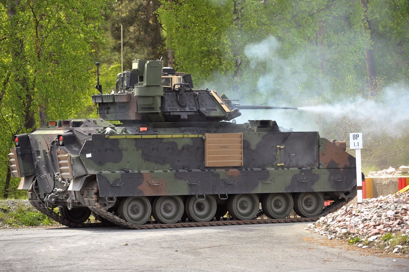 A Bradley fighting vehicle crew operates their vehicle on a firing range at the Grafenwoehr Training Area in Germany during the Combined Resolve II exercise in 2014. The photo shows the Bradley positioned on a training lane with its turret oriented toward target areas. Crew members, partially visible through open hatches, are focused on gunnery tasks and target engagement. The background includes the open, rolling terrain and forested areas typical of Grafenwoehr. This image illustrates how U.S. Army teams train for precision and speed in weapons operation. The exercise brought together soldiers from multiple NATO nations to develop skills and interoperability. The M2 Bradley's role as a fighting vehicle for mechanized infantry is highlighted in this training environment.