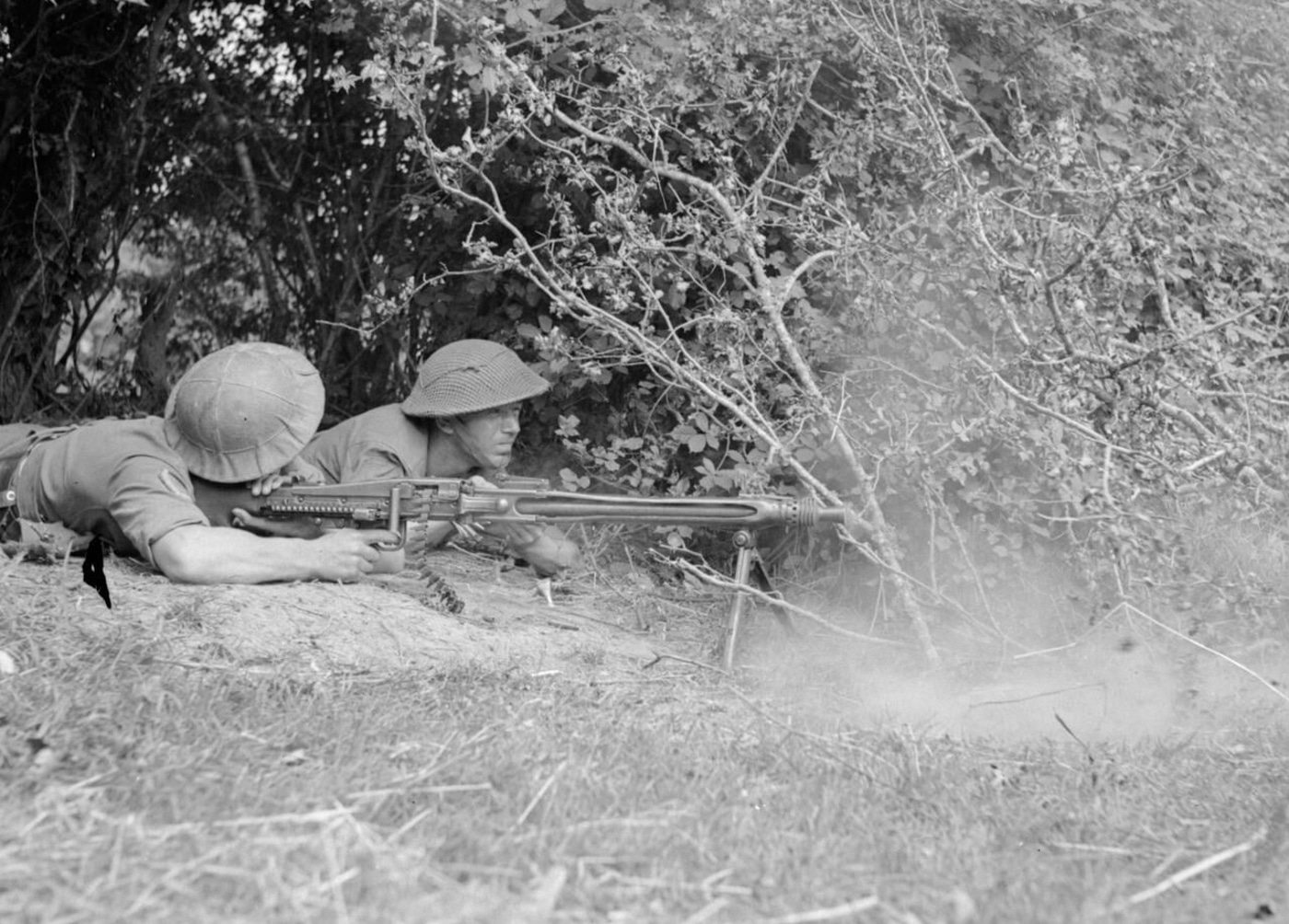 British soldiers are pictured operating an MG 42 machine gun during a non-commissioned officer (NCO) training course. One soldier is prone behind the weapon, firing it, while another assists by feeding the ammunition belt into the MG 42. The men wear standard British uniforms and helmets, and the scene is set outdoors in a training area, with dirt and sparse grass underfoot. The MG 42’s bipod, receiver, and long ammunition belt are clearly visible. This photo shows Allied troops learning to handle and understand captured German machine guns as part of their NCO school curriculum.