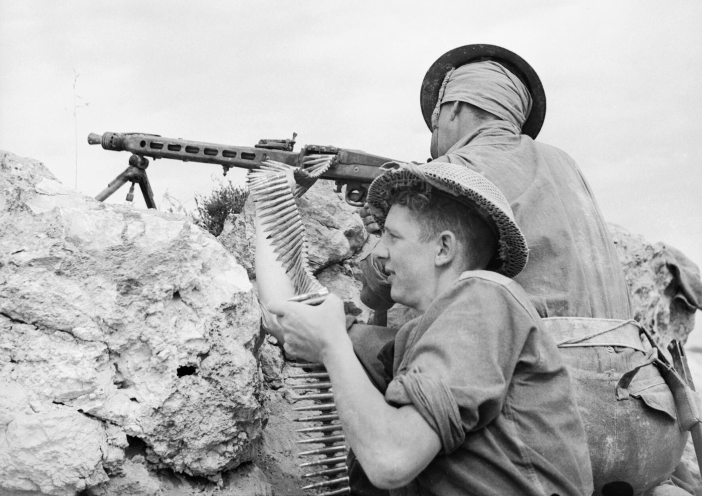 British soldiers from the 2nd Sherwood Foresters are seen firing a captured German MG 42 machine gun on April 27, 1943. The men are positioned on the ground, working together to operate the unfamiliar weapon, with one man on the trigger and another feeding the ammunition belt. Their British uniforms, webbing, and helmets contrast with the German machine gun at the center of the scene. The MG 42 is set on its bipod, and spent casings are scattered on the ground nearby. This photo highlights Allied troops testing and using captured enemy weapons during World War II. The expressions and posture of the men suggest focused effort and curiosity about the MG 42’s performance.