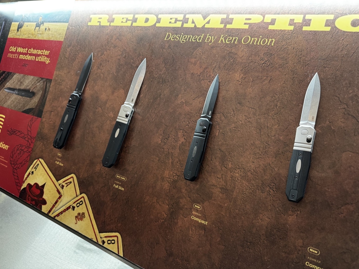 This image shows the CRKT Redemption Series knives arranged on a display table at the 2025 Blade Show. Three models are visible: a full-size all-black version and two compact versions — one blacked out and one with a traditional finish. All knives are laid out side by side with their MagnaCut blades open, revealing the unique coffin-handled shapes inspired by Ken Onion’s Deadman’s Hand customs. Product tags or information cards are visible near each knife, and the CRKT booth’s branded table covering sets the scene. The lighting accentuates the finishes on the knives, from the matte black Cerakote to the polished edges, while the modern, tactical styling is clear. This focused display lets attendees compare the Redemption models up close.