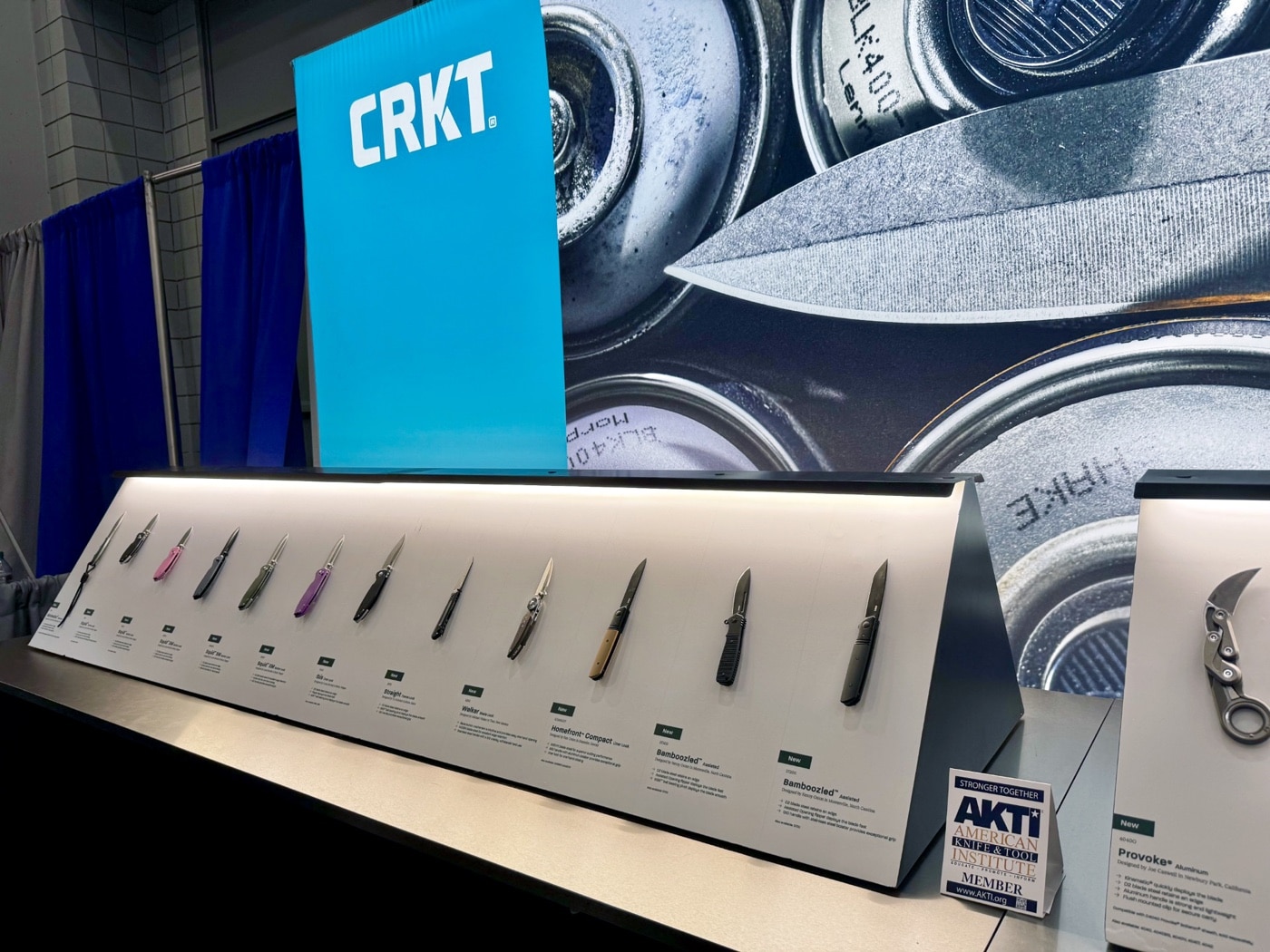 This image shows the CRKT (Columbia River Knife and Tool) display booth at the 2025 Blade Show in Atlanta. Several knives are arranged on branded display tables, including the Redemption Series designed by Ken Onion. Attendees can be seen examining the knives, picking them up, or talking with CRKT representatives. Promotional banners and product cards are visible in the background, featuring the CRKT logo and marketing messages. The booth is well-lit, and the layout is neat and organized, with a focus on new knife models and collaborations. The atmosphere is busy but inviting, reflecting the brand’s active presence at the show.