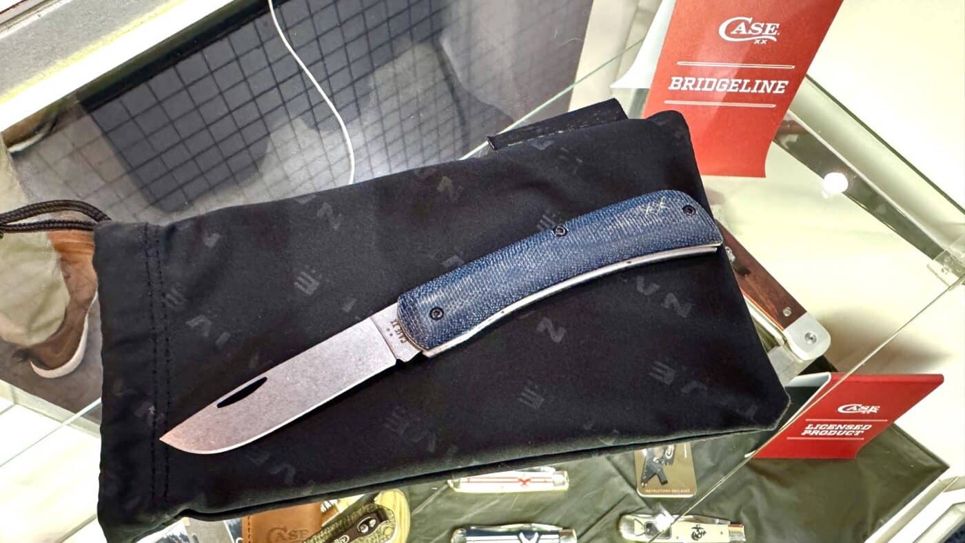 This image features the Case Crossroads slip joint knife fully open and resting on top of a glass display case at the 2025 Blade Show. The knife’s drop point blade is made from stone-washed S35VN stainless steel and is positioned to show its flat grind and clean edge. The handles are made from smooth blue denim canvas laminate with white G-10 liners, offering a modern, eye-catching appearance. Reflections from the display case glass subtly highlight the contours of the knife. Other Case products or show items are visible inside the case below, but the Crossroads is the primary focus of the photo. The lighting inside the convention hall emphasizes the blue tones of the handle and the finish of the blade. This close-up shot allows viewers to see the details and craftsmanship of Case’s new model.