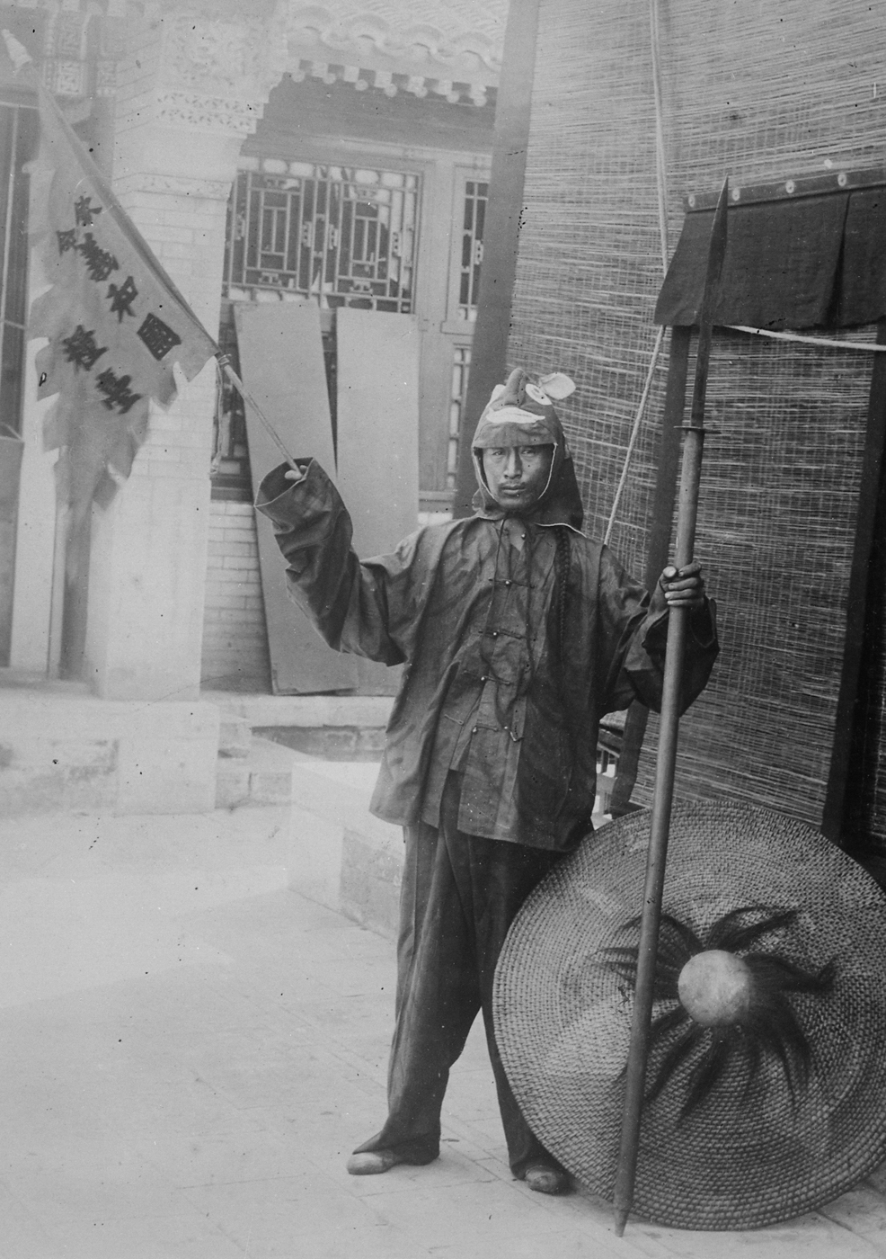 A Chinese Boxer rebel, identified by his simple rural clothing and martial arts stance, stands in a street or courtyard in Peking (Beijing) holding a distinctive flag. The flag may be inscribed with Chinese characters or traditional symbols linked to the Society of the Righteous and Harmonious Fists, the group leading the Boxer Rebellion. The young man’s expression is serious and determined, reflecting the passion and anger of those involved in the uprising. Behind him, architectural details like brick walls, wooden gates, or tiled roofs hint at the urban setting of Peking during 1900. The image highlights the grassroots nature of the Boxer movement and the strong anti-foreign sentiment that fueled their cause. His presence with the flag shows how symbolism and ritual were central to rallying support and intimidating both Chinese converts and foreigners. This photo represents the human face of the Boxer Rebellion amid the chaos and violence of turn-of-the-century China.