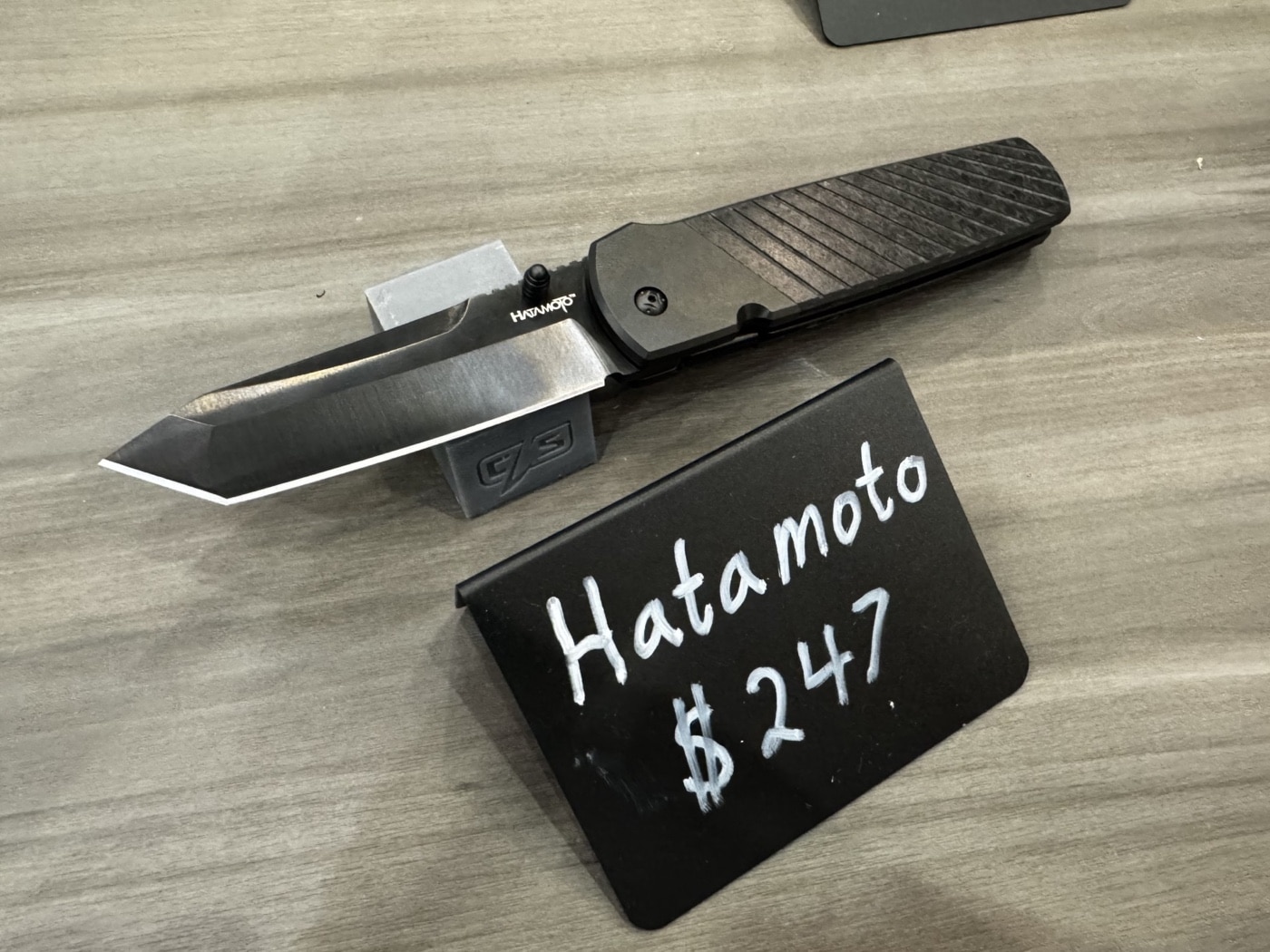 This image shows the Cold Steel Hatamoto folding knife on display at the Cold Steel booth during the 2025 Blade Show. The knife is laid out with the blade open, revealing a 4-inch tanto blade made from CPM 20CV steel, which has a polished finish. The handle is crafted from full carbon fiber, giving it a lightweight yet high-end look. The knife rests on a branded display surface, and there may be a product card or label nearby with specifications. Other Cold Steel products and booth signage are visible in the background, but the Hatamoto is the clear focus of the shot. Lighting from the convention floor highlights the blade’s shape and the texture of the carbon fiber handle.