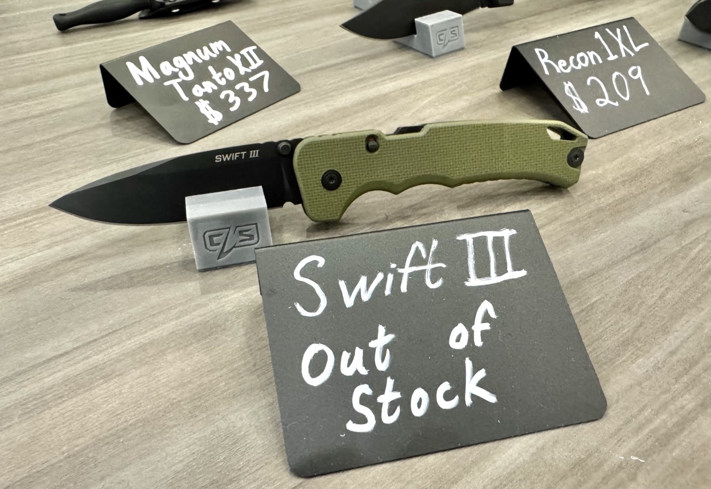 This photo features the Cold Steel Swift III knife displayed open at the Cold Steel booth during the 2025 Blade Show. The knife’s 4-inch CPM M4 tool steel blade is extended, showcasing a drop point shape with a stonewashed finish. The handle is made of textured black G10, and the unique Atlas lock mechanism is visible near the pivot. The knife is set on a branded mat or display surface, with product cards or specs possibly placed nearby. Cold Steel signage and other knives are visible in the background, but the Swift III is in clear focus. The lighting highlights the blade’s grind and the handle’s texture, allowing viewers to see key details of this popular assisted-opening model.