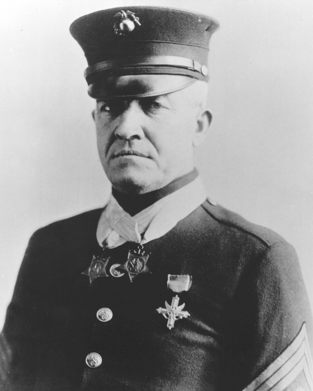A formal studio portrait of Dan Daly, wearing his U.S. Marine Corps uniform with two Medals of Honor prominently pinned to his chest. Daly’s expression is composed and serious, reflecting his status as one of America’s most highly decorated Marines. The double medals are clearly visible, each suspended from its ribbon, signifying extraordinary bravery in combat. The photograph’s background is neutral, ensuring full focus on Daly and his achievements. His uniform includes service stripes and other decorations, and his posture is upright, displaying both pride and humility. The image is black and white or sepia-toned, typical of early 20th-century military portraits. This photo is often used to illustrate the legacy of Dan Daly’s heroism during the Boxer Rebellion and World War I, emphasizing his rare place in military history.