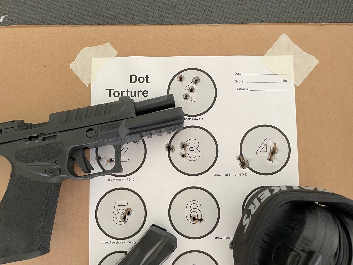 The photo shows a Springfield Echelon pistol placed next to a Dot Torture Drill target, both set on a bench at a public shooting range. The Dot Torture target is an 8.5”x11” sheet with ten numbered dots, each with visible bullet holes from a recent shooting drill. The Echelon is unloaded and positioned for safety, following standard range procedures. The image highlights the connection between effective pistol training and tracking progress with well-known drills. Public range features like individual shooting lanes and safety signage are visible in the background. The combination of this modern pistol and the Dot Torture Drill underscores the importance of accuracy, trigger control, and consistency. The photo supports content about skill development, safe practice routines, and using practical drills at public ranges.