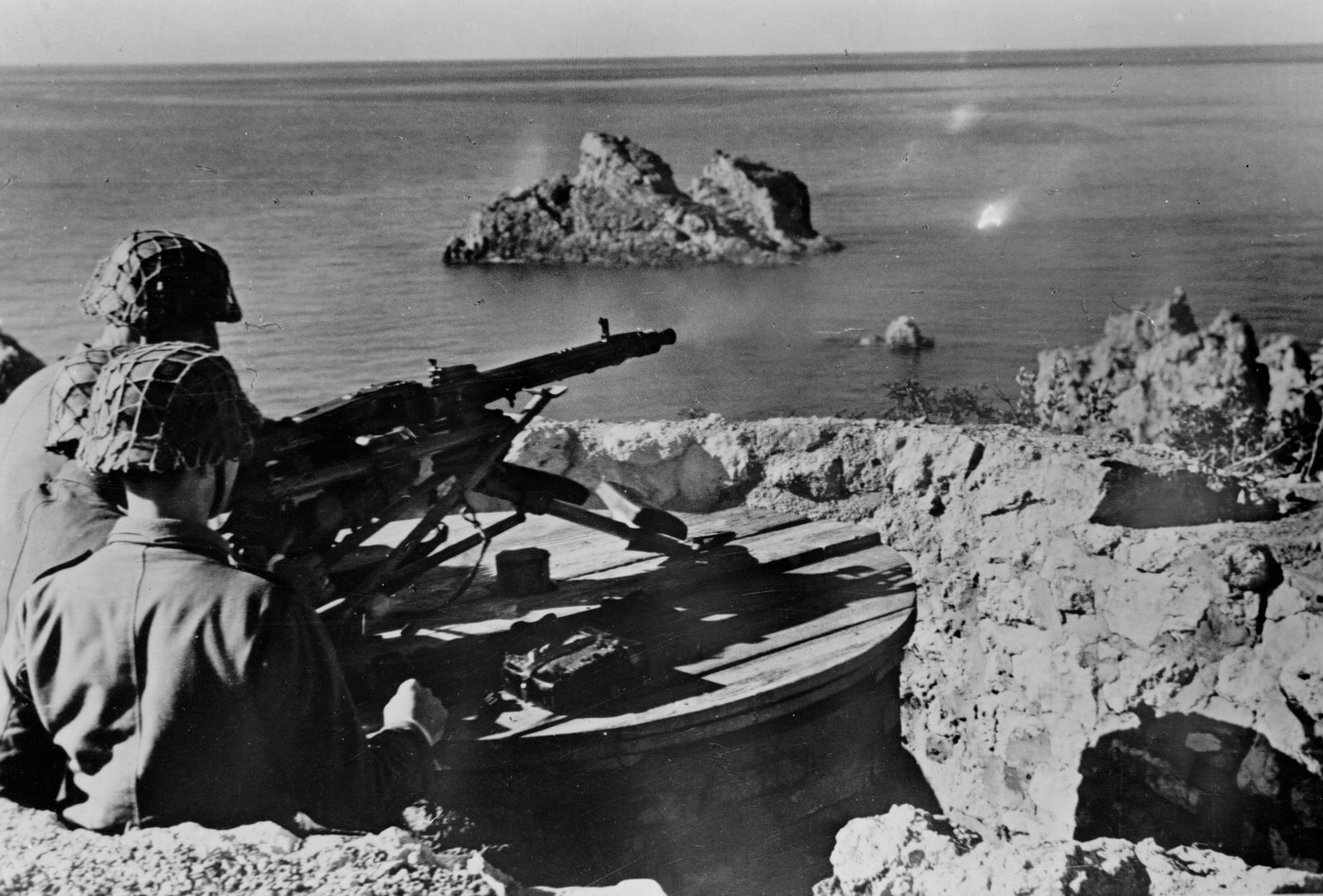 A German MG 42 machine gun is set up in a defensive position overlooking the coastline of the Greek island of Corfu, with the Ionian Sea visible in the background. Soldiers from the 4th SS Panzergrenadier Division man the position, some crouched behind sandbags or rocks for cover. The MG 42 sits on its bipod, with ammunition belts nearby and field gear scattered around. The soldiers wear typical SS uniforms and helmets, focused on scanning the shoreline for enemy activity. The photo highlights the strategic use of the MG 42 machine gun in coastal defenses during World War II.