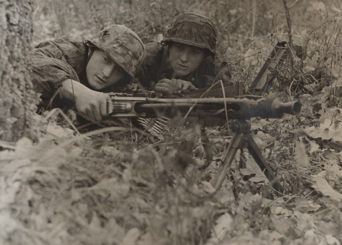 German SS armored grenadiers are crouched behind makeshift cover with an MG 42 machine gun set up in a defensive position during the Battle of Korsun-Cherkassy bridgehead in World War II. The gun crew is focused on their sector, with ammunition belts visible and one soldier appearing to ready the MG 42 for firing. Their uniforms are worn and their gear includes steel helmets and field equipment typical of SS troops in 1944. The MG 42 machine gun, known for its high rate of fire and distinctive sound, is at the center of the photo. Background shows a bleak, war-torn landscape with scattered debris and muddy ground. This image highlights the German use of the MG 42 in major Eastern Front battles.