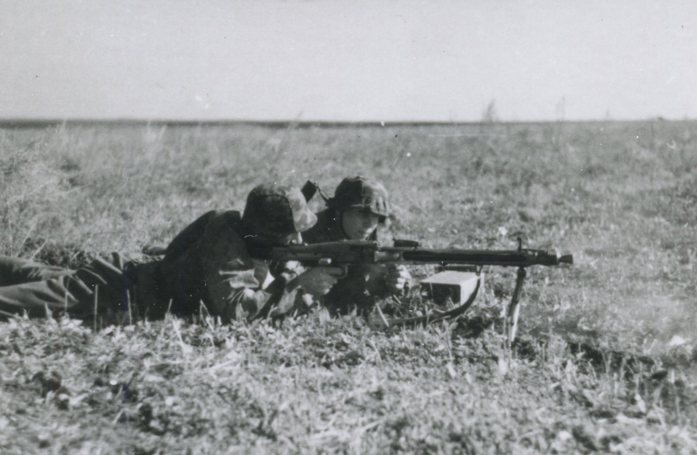 German SS soldiers are lying prone in tall grass on the Eastern Front, firing an MG 42 machine gun. One trooper mans the weapon while another assists, feeding a long belt of 7.92mm ammunition. Both wear standard issue field gear, including helmets and camouflage smocks. Spent shell casings and disturbed earth mark their firing position. The MG 42’s distinctive shape and bipod are clearly visible, highlighting its role as the primary squad automatic weapon. This scene shows typical combat conditions for German forces using the MG 42 machine gun against Soviet troops.