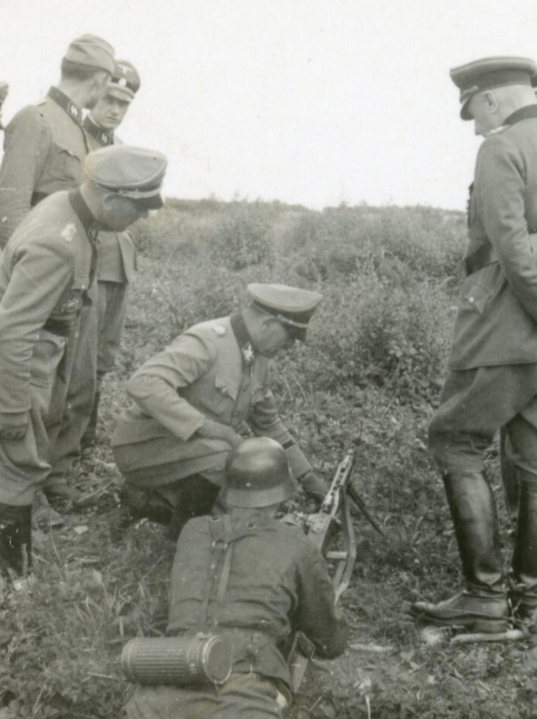 A group of German officers observes a demonstration of the newly introduced MG 42 machine gun. One soldier operates the MG 42, either firing or explaining its features, while the officers watch closely, some with hands behind their backs or taking notes. The MG 42 is set up on its bipod and aimed downrange, with visible ammunition belts prepared for use. The officers’ uniforms suggest a mix of field and administrative roles, and their body language shows interest in the weapon’s operation. The scene emphasizes how the German Army introduced the MG 42 machine gun to its leadership through practical demonstrations and briefings.