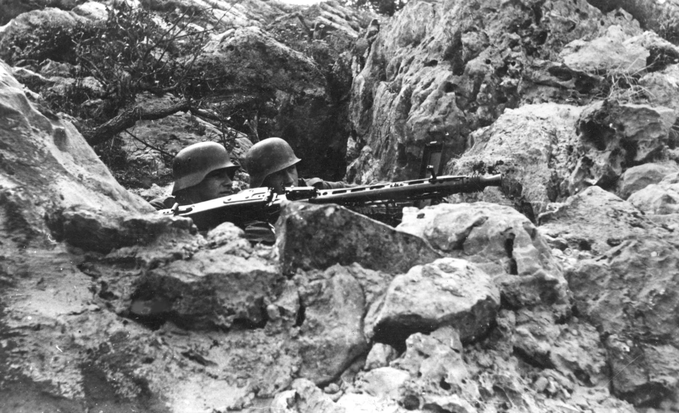 An MG 42 heavy machine gun is set up in a fortified position among rocky terrain on the Italian front in 1944. The gun sits on its bipod, aimed down a mountain pass, with sandbags and rocks used for extra cover. Several German soldiers, partially visible, are positioned nearby — one on the weapon and others watching for enemy movement. The background shows rugged mountain slopes and sparse vegetation. The scene highlights the challenges of using the MG 42 machine gun in harsh, elevated conditions typical of the Italian campaign. Details like ammunition boxes and field gear add to the sense of a prepared defensive post.