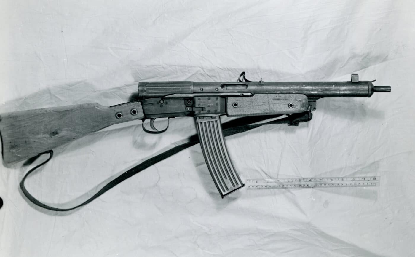 A black-and-white archival photograph showing a single Gustloff Volkssturmgewehr rifle being inspected on a workbench by US Ordnance technical intelligence in late May 1945. The image captures the crude, utilitarian design features of the weapon, including its stamped metal receiver, fixed wooden stock, and ribbed magazine. The rifle is laid out for detailed examination by American personnel, who are visible in the background taking notes or handling technical documents. Lighting in the photo is harsh, emphasizing the rough surfaces and minimal finishing typical of Volkssturmgewehr production. This review process took place immediately after the end of World War II in Europe, as the Allies evaluated captured German arms. The context and time period are critical for understanding the historical significance of the rifle. The photo documents one of the few surviving examples of a late-war German Volkssturmgewehr rifle, highlighting its role in the final months of the conflict.