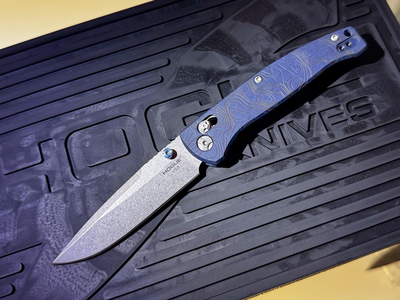 This image shows the Hogue Mysto Water folding knife on display at the Hogue booth during Blade Show 2025, with the blade open. The blade is crafted from CPM MagnaCut stainless steel and has a stone-tumbled finish, emphasizing its fine edge and clip point profile. The handle is made of blue Richlite with laser-engraved topographic details representing the Central California seafloor, giving it a unique oceanic appearance. The knife is positioned on a branded display surface, with Hogue booth materials and product literature nearby. Bright show lighting enhances the blue tones and engraved patterns on the handle, and the knife’s light weight and slim construction are visible. The photo highlights Hogue’s craftsmanship and the Mysto Water’s modern features.