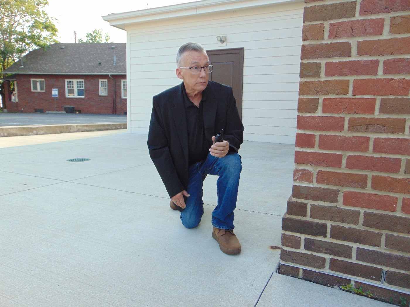 The photo shows the article’s author in a kneeling position, wearing a dark 5.11 Founder’s Jacket and 5.11 Chukka Boots. While the jacket looks like an ordinary sport coat, it discreetly conceals a full complement of safety equipment, including a CCW handgun and a Taser, both carried securely inside the jacket’s internal pockets and loops. The author’s posture indicates readiness and comfort, demonstrating that movement is not restricted by the gear. The boots look sturdy and blend seamlessly with casual or business-casual clothing. This image illustrates the effectiveness of the Founder’s Jacket for security professionals or armed civilians who need to carry multiple tools without drawing attention. The focus is on the versatility and concealment features of the 5.11 apparel in an active setting.