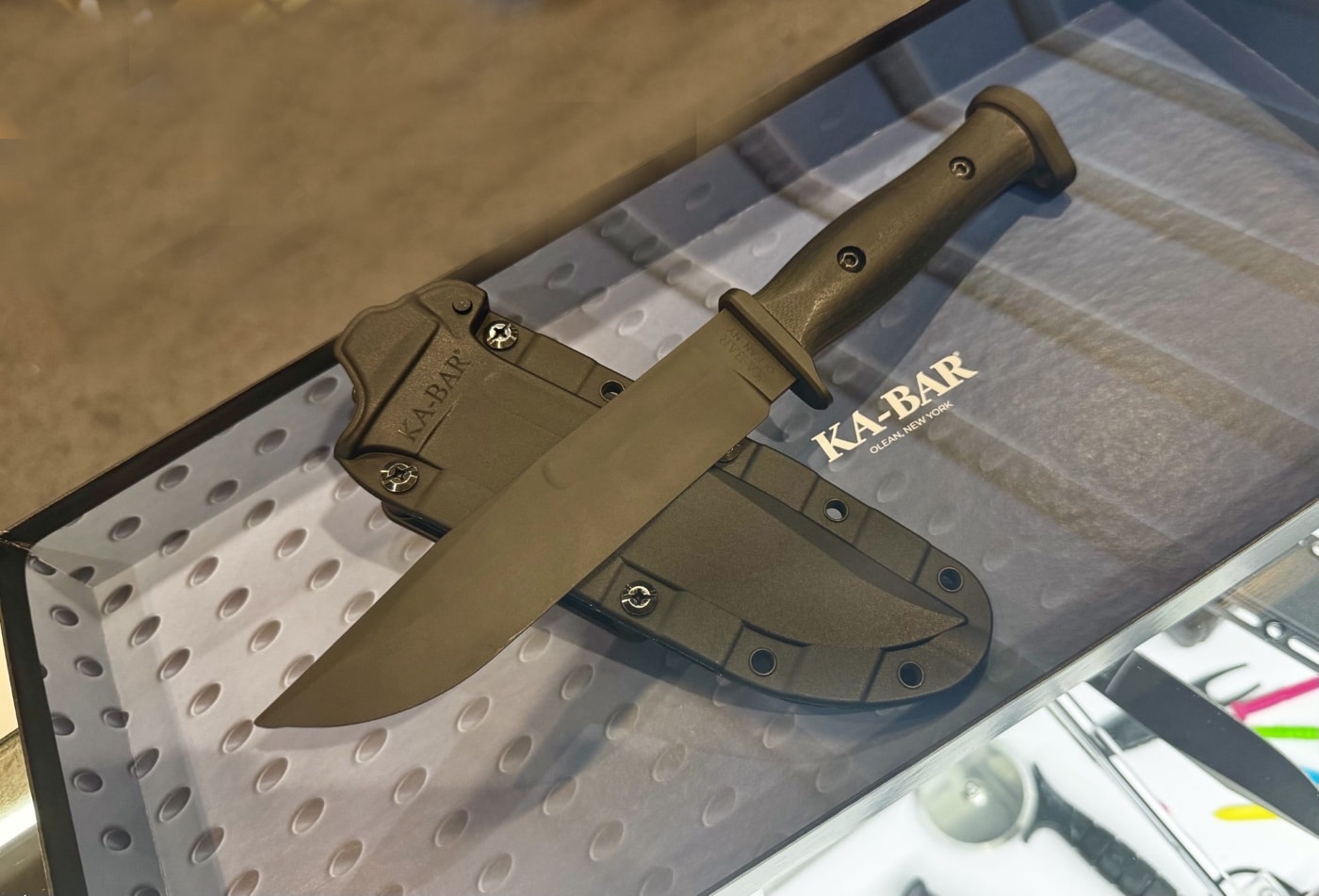 This image provides a close-up view of the Ka-Bar “Slabby” fixed blade knife during a sneak peek at the 2025 Blade Show. The knife is displayed with its blade exposed, showing a coated 1095 steel construction and a wide, full tang profile. The handle features large, flat scales — likely removable or interchangeable — for enhanced grip and customization. The knife rests next to its injection-molded sheath, which includes a double locking mechanism and multiple mounting points. The Ka-Bar booth setup, with other knives and branding materials, is visible in the background. The overall look is modern and utilitarian, highlighting both the secure sheath and the robust build of the Slabby.