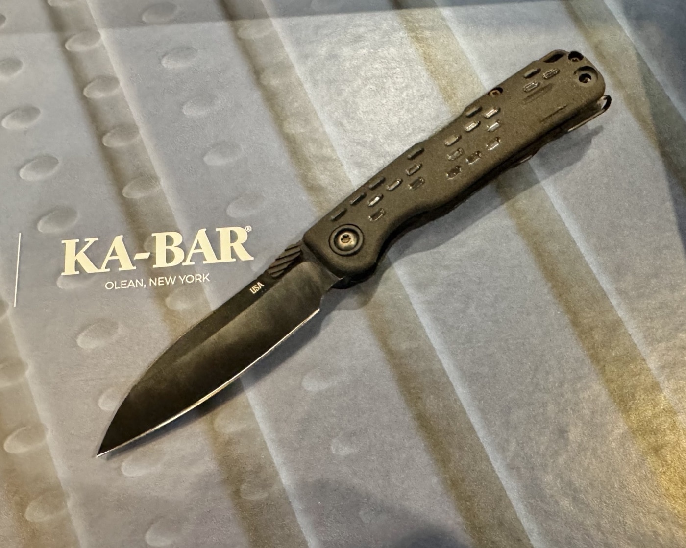 This image shows a Ka-Bar representative handing the State & Union folding knife to the author at the 2025 Blade Show. The knife is open, displaying its drop point blade made from German 1095 Cro-Van steel with a black oxide coating. The handle is constructed from fiberglass-reinforced nylon (FRN), offering a textured and lightweight grip. The author holds the knife while listening to the rep’s explanation, and the booth’s branded materials, display stands, and informational cards are visible in the background. The interaction takes place in a well-lit section of the trade show floor, surrounded by other Ka-Bar products. The image highlights the knife’s accessible design and the hands-on experience typical of the Blade Show.