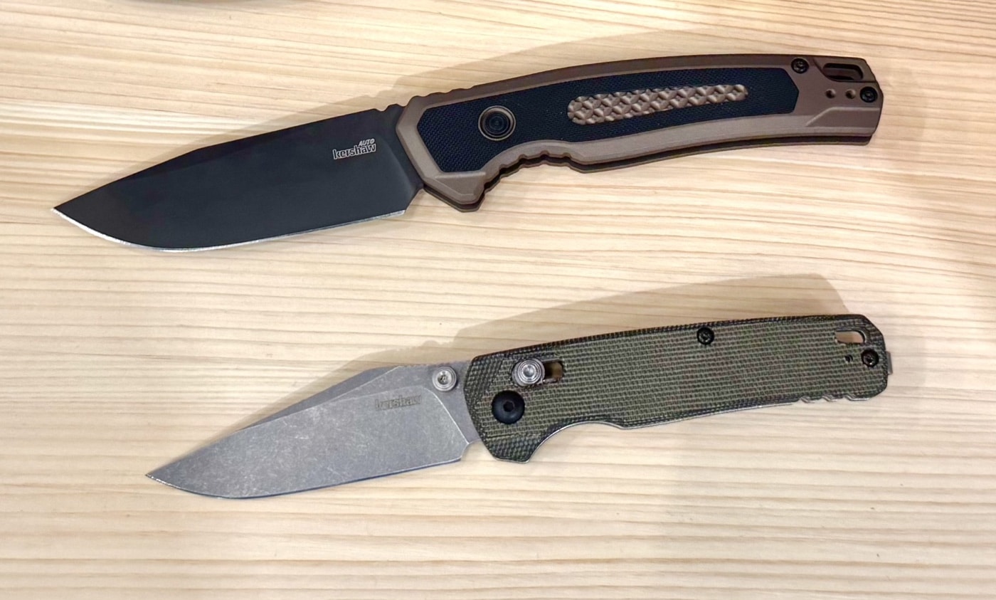 This image features two Kershaw knives displayed on a table at the 2025 Blade Show, with the Launch 21 automatic folder positioned at the top and the Bel Air clip point knife placed directly below. The Launch 21 has a flat dark earth anodized aluminum handle with black G10 inserts and a drop point CPM M4 blade with a black PVD coating. The Bel Air, beneath it, features olive canvas micarta handle scales and a CPM MagnaCut blade in a working finish. Both knives are shown open, highlighting their blade shapes, handle textures, and overall profiles. Branded display mats and Kershaw product cards may be present in the background, while booth lighting brings out the color contrasts and finishes. The arrangement makes it easy to compare these two new models side by side.