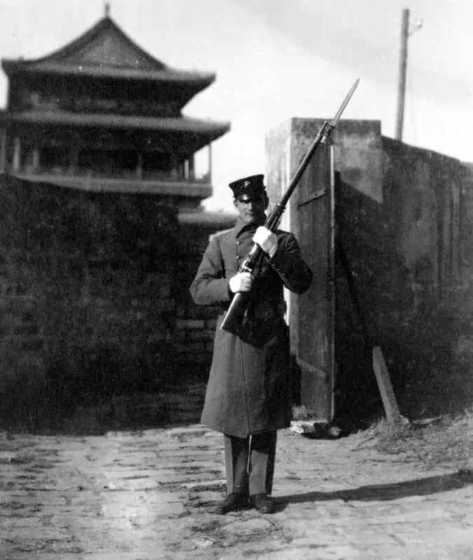 A U.S. Marine, dressed in early 1900s field uniform, stands at his post gripping a Krag–Jørgensen rifle with both hands. The Marine’s stance is upright and alert, with the bolt-action rifle held diagonally across his chest or at his side, ready for immediate use. The details of the Krag rifle are clear, including its distinctive side loading gate, wood stock, and metal barrel. The Marine’s uniform features a campaign hat, cartridge belt, and simple field gear, emphasizing the period authenticity. Background elements may include sandbags, a low wall, or basic camp structures, suggesting a defensive or outpost setting. The image highlights the standard weaponry used by U.S. Marines during the Boxer Rebellion and underlines the importance of the Krag–Jørgensen in American military history. The photo provides context for how Marines were equipped and the conditions they faced during overseas operations in China.