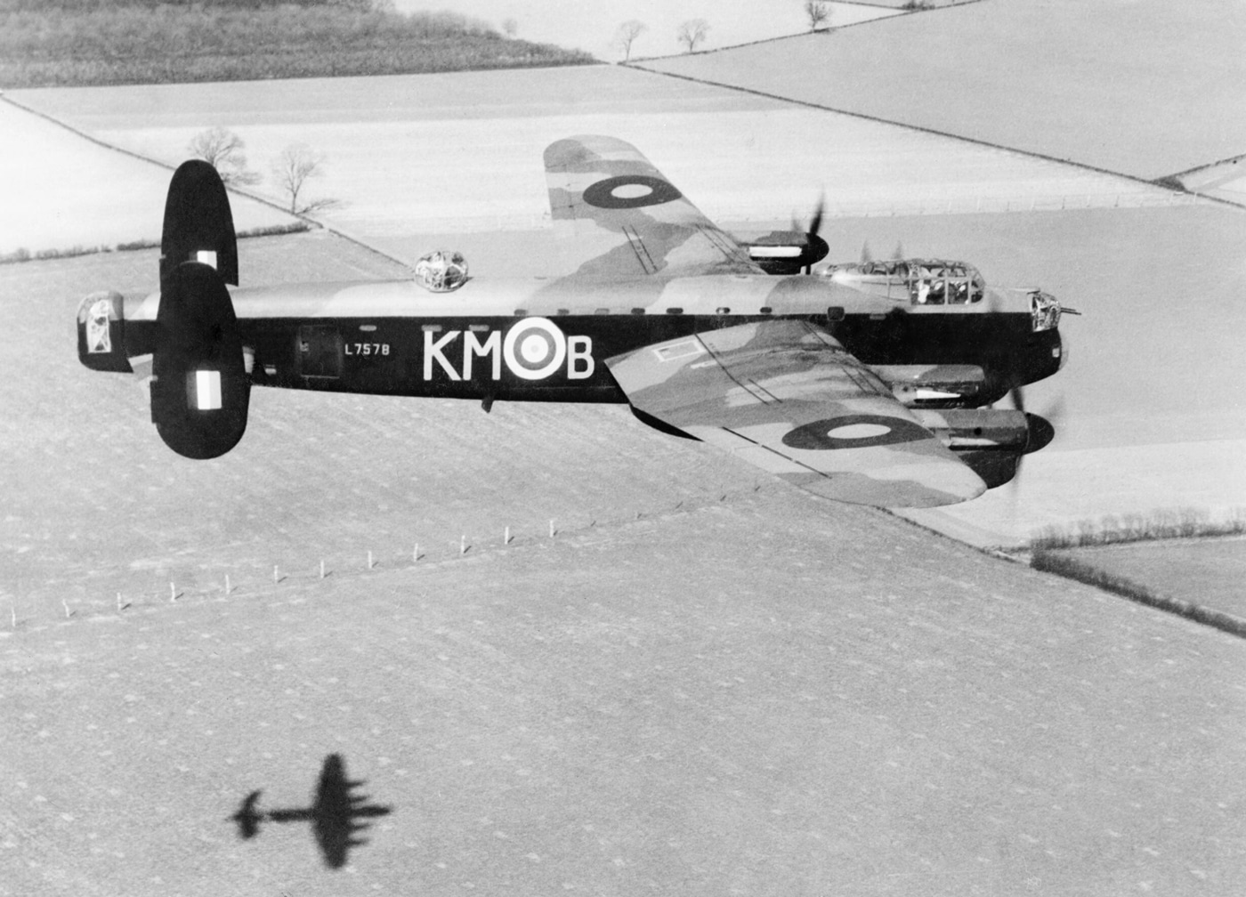 The photograph shows Lancaster Mark I, L7578, code KM-B, belonging to No.44 (Rhodesia) Squadron, Royal Air Force, flying at low altitude over the Lincolnshire countryside on 14 April 1942. The bomber is seen from the side or slightly below, with its four engines and characteristic twin tailfins clearly visible against a backdrop of green fields and hedgerows. The aircraft is in full operational camouflage and marked with RAF roundels and squadron codes, preparing for the upcoming low-level attack on the M.A.N. diesel engineering works at Augsburg. The low altitude of the flight is evident from the visible detail in the English landscape beneath the bomber. This scene is part of the intensive training undertaken by RAF crews before launching one of the war’s most daring daylight raids. The image captures both the technical features of the Lancaster and the context of pre-mission practice flights over rural England. There are no other aircraft in the frame, emphasizing L7578’s solo training sortie.