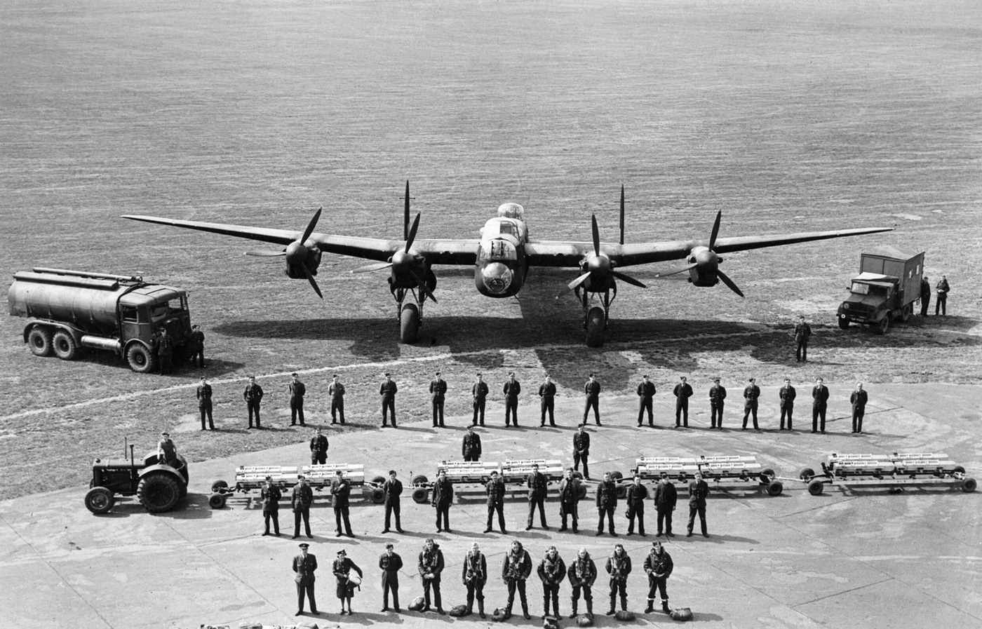 The photograph displays a comprehensive line-up of all RAF Bomber Command personnel necessary to keep a single Avro Lancaster heavy bomber operational during wartime missions. The image shows the full range of support staff and aircrew, including pilots, navigators, engineers, armorers, mechanics, radio operators, and administrative staff, all standing in front of or beside the Lancaster. The bomber itself is visible in the background, providing context for the scale of manpower required for maintenance, arming, fueling, and preparation for each mission. The personnel are dressed in a mix of flight suits, service uniforms, and work attire, indicating their different roles and responsibilities. The formation stretches along the length of the aircraft, emphasizing the teamwork and coordination needed to ensure safe and effective operations. The photo gives a clear visual impression of the logistics behind every successful sortie, not just the actions of the flight crew. The setting is likely an airfield or dispersal area typical of WWII RAF bomber bases.