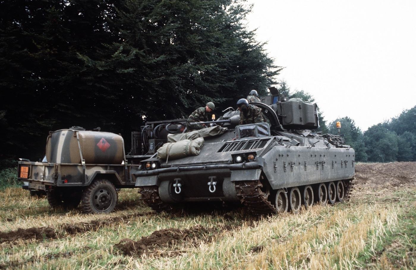 This image shows a U.S. Army M2 Bradley infantry fighting vehicle and its crew preparing to advance through the German countryside during Exercise REFORGER ’85. The Bradley is positioned in a grassy or lightly wooded rural area, with its turret facing forward and soldiers gathered nearby or inside the vehicle. Crew members in combat uniforms and helmets are making final checks or discussing their route before moving out. The surrounding landscape is typical of West Germany, featuring open fields, rolling terrain, and perhaps distant farmhouses or trees. The scene reflects the focus on readiness, coordination, and mobility that defined Cold War-era NATO training exercises. The Bradley’s armored hull, side skirts, and mounted weapons are clearly visible. This type of field exercise was central to ensuring U.S. forces could quickly respond to any threat in Europe.