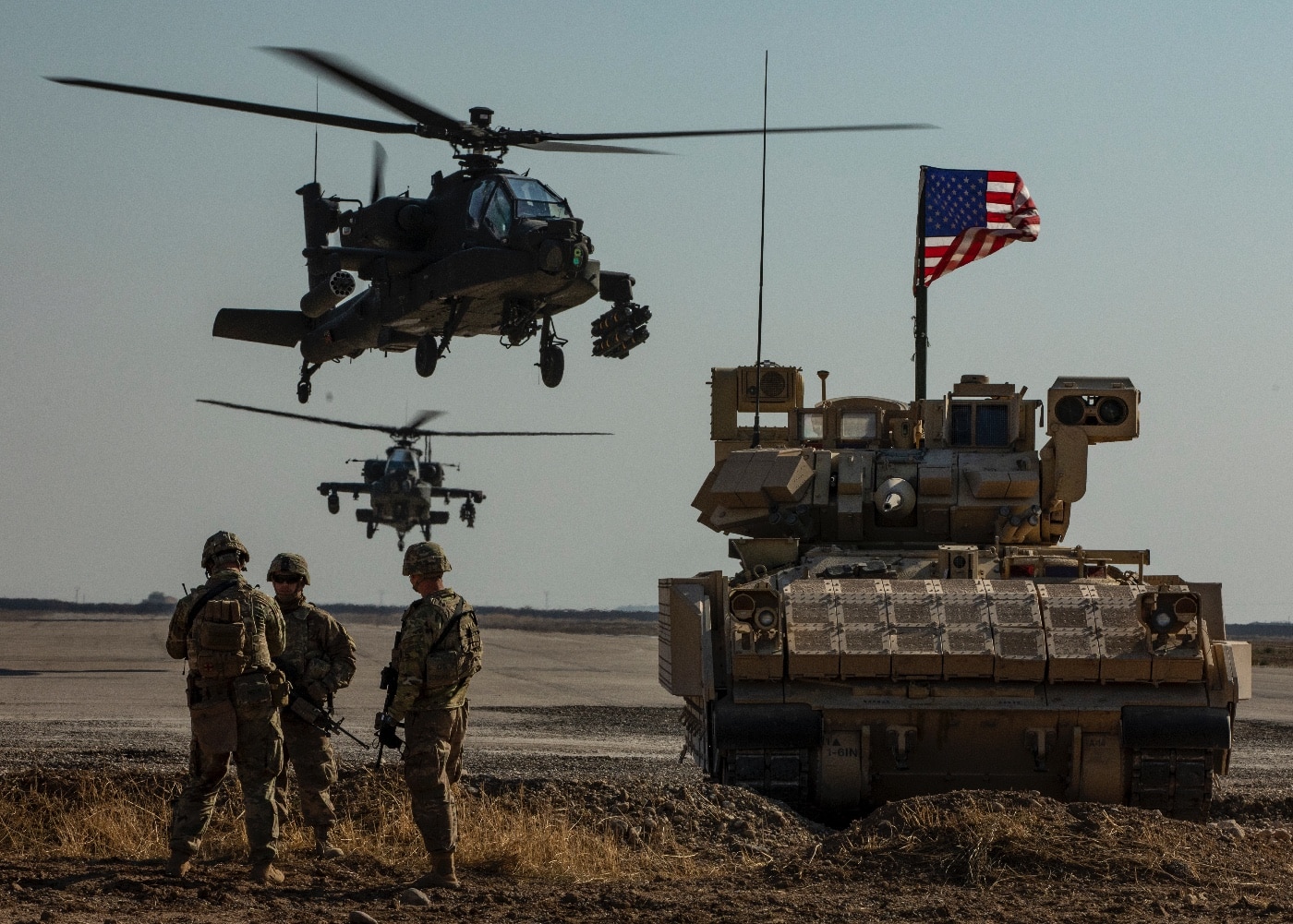The image features soldiers from the U.S. Army’s 1st Armored Division posing for a photo with an M2 Bradley infantry fighting vehicle and two AH-64 Apache attack helicopters in the background. The scene takes place in a desert area in Syria, with sandy ground and clear skies typical of the region. The Bradley is positioned at the center, while the Apaches are parked nearby, showcasing both ground and air assets used in Operation Inherent Resolve. The soldiers are wearing full combat uniforms, some holding rifles, and standing or kneeling in front of the vehicle. This image highlights the integration of armored vehicles and aviation in joint operations. The setting underlines the logistical and operational reach of U.S. forces in deployed environments. The photo captures a sense of camaraderie and professionalism among the unit members.