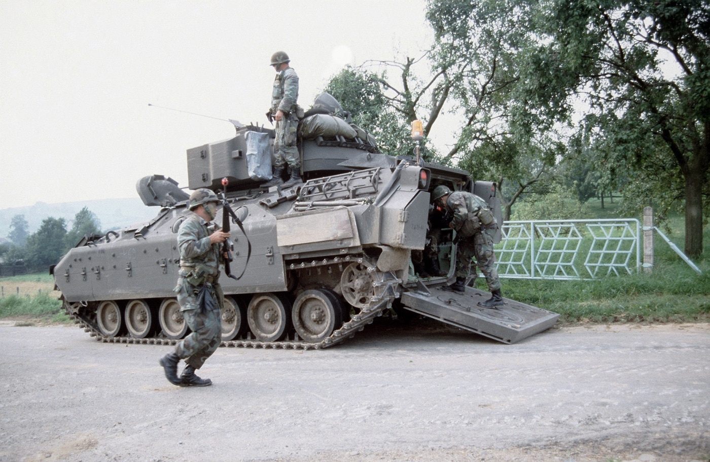 This image shows the right side of a U.S. Army M2 Bradley infantry fighting vehicle stopped along a paved or dirt road during exercise REFORGER ’85. The Bradley’s side armor, road wheels, and turret are clearly visible, with the vehicle’s camouflage pattern blending into the European surroundings. The vehicle is at rest, positioned parallel to the road, with no crew visible outside. The background includes trees or brush typical of Central European training areas, and the sky is either overcast or partly cloudy. The scene emphasizes the vehicle’s profile and mobility features, such as its track system and side-mounted stowage. REFORGER exercises focused on rapid deployment and large-scale maneuvers, preparing U.S. and NATO forces for potential conflict scenarios. The image provides a straightforward view of the M2 Bradley’s design and use in 1980s-era training environments.