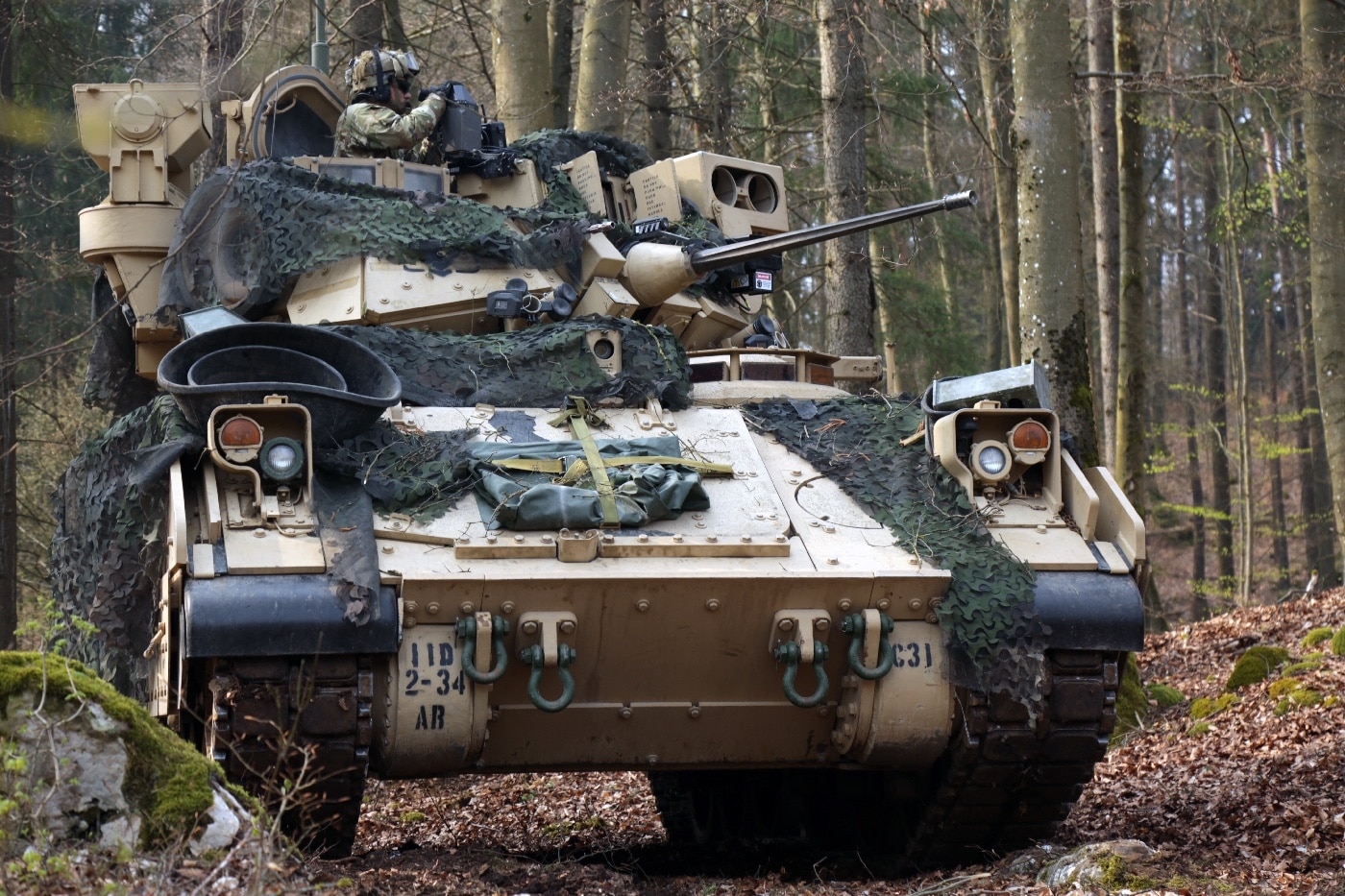 A U.S. Soldier with the 34th Armored Regiment is pictured standing inside a Bradley fighting vehicle, scanning the surrounding area during the Allied Spirit X military exercise held in Hohenfels, Germany, in 2019. The vehicle's turret and observation equipment are visible, emphasizing the Bradley’s role as both transport and fire support on the battlefield. The soldier is wearing standard combat gear and helmet, focusing intently on their sector. The surrounding terrain includes open ground and sparse vegetation typical of European training ranges. This image highlights the operational readiness and crew vigilance that the M2 Bradley supports. It also demonstrates U.S. and NATO collaboration during multinational training. The M2 Bradley, America’s primary infantry fighting vehicle, is central to these types of exercises.