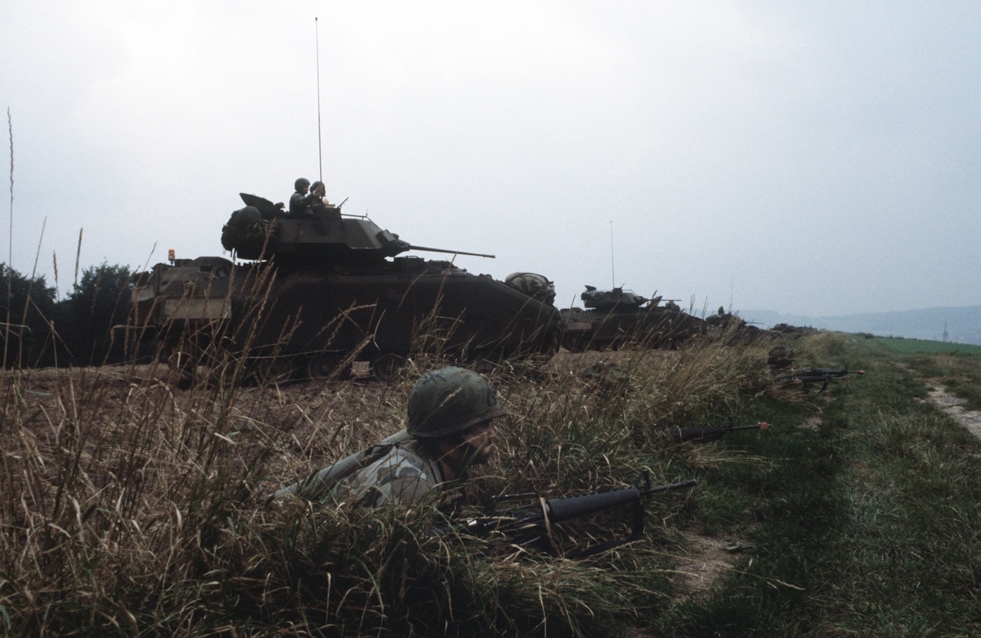 The image shows three U.S. Army M2 Bradley infantry fighting vehicles advancing in single file through a wooded area during Exercise REFORGER ’85. The vehicles are spaced out for tactical movement, navigating between trees and brush in typical Central European forest terrain. The Bradleys’ angular shapes and distinctive turrets are visible, with camouflage paint blending into the green and brown environment. Their tracks leave clear marks in the soft forest floor as they maneuver. This exercise, held in 1985, was part of NATO’s efforts to test large-scale mobility and coordination during the Cold War. The photo captures the challenges of operating armored vehicles in restricted and rugged natural terrain. M2 Bradleys were designed to support infantry while moving rapidly through such environments.