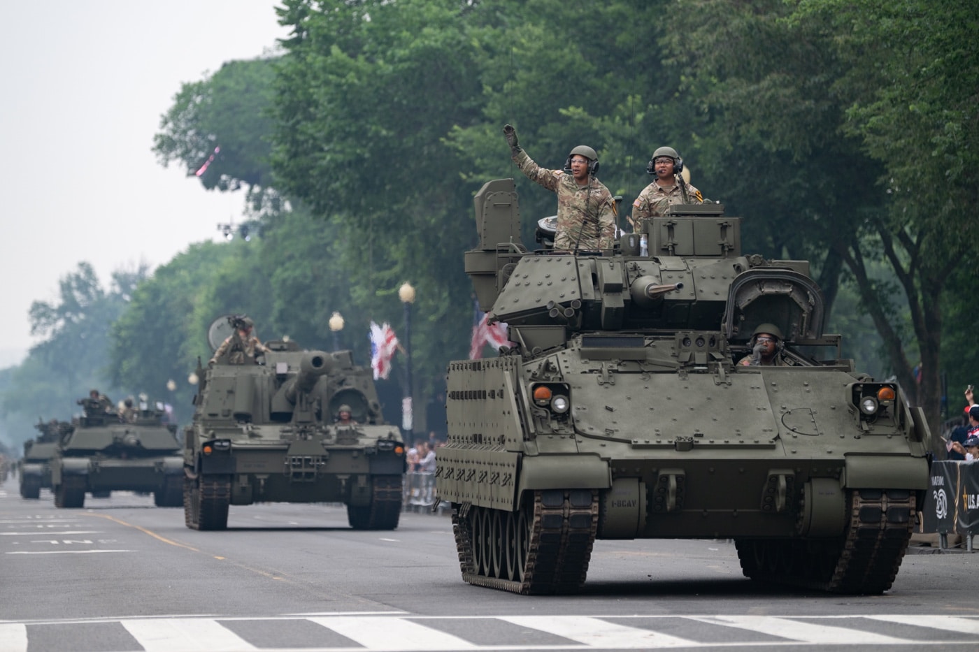 The image captures a U.S. Army M2 Bradley infantry fighting vehicle rolling along Constitution Avenue in Washington, D.C., as part of the 250th Army Birthday Parade on June 14, 2025. Soldiers in combat gear ride inside or behind the vehicle, maintaining disciplined formation. In the background, viewers line the streets and federal buildings along the parade route. The scene combines ceremonial celebration with military hardware, reflecting both history and readiness. The Bradley’s turret and armored side are prominently visible, positioned as part of a broader column of vehicles including M1 Abrams tanks and Strykers.