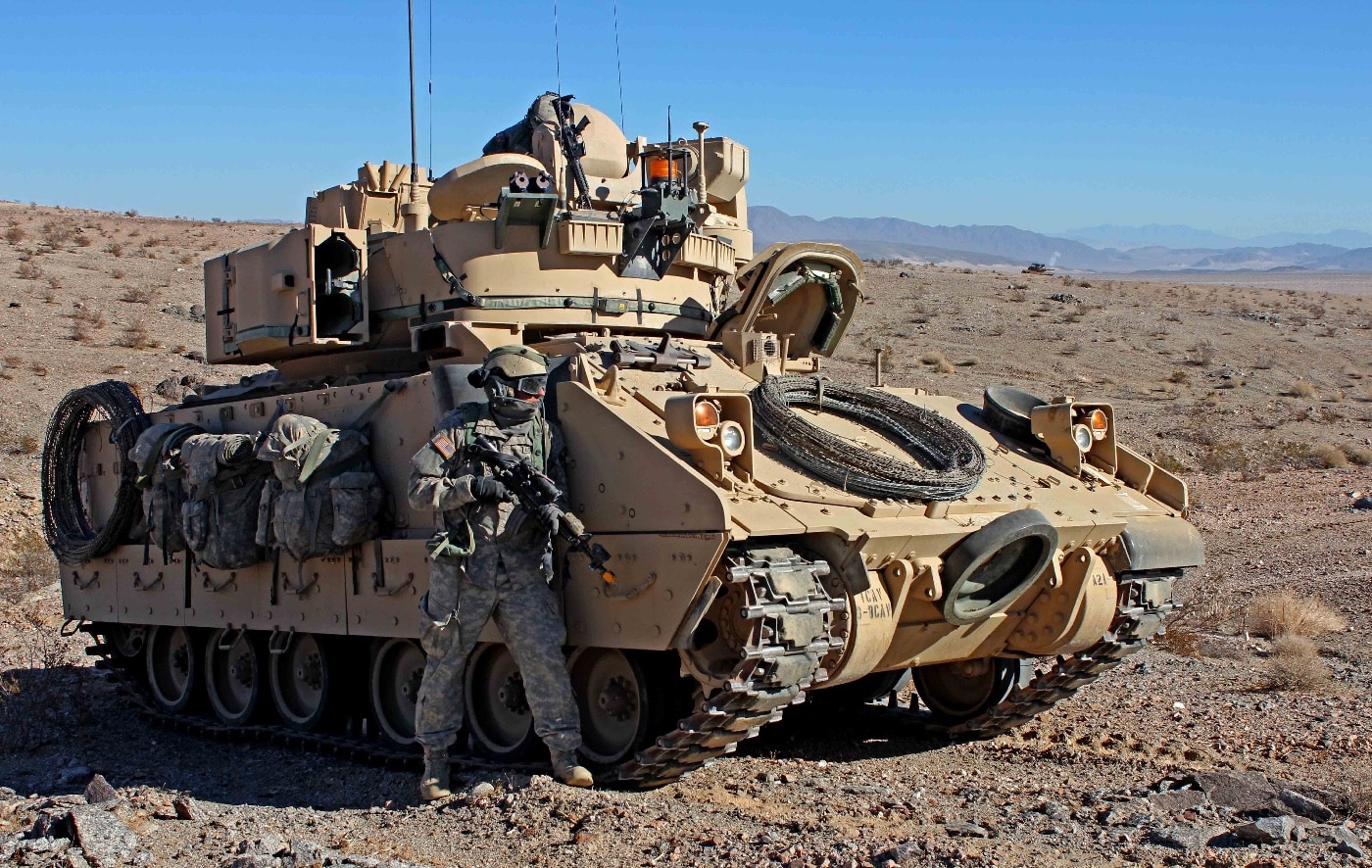 This image shows a U.S. Army soldier from the 1st Infantry Division pulling security next to an M2 Bradley infantry fighting vehicle at the National Training Center in Fort Irwin, California. The soldier is in full combat gear, holding a rifle and positioned in a low, alert stance near the vehicle’s side. The Bradley is parked on arid, sandy ground typical of the Mojave Desert, with sparse vegetation and distant hills visible in the background. The soldier’s focus is outward, scanning the environment for potential threats during the exercise. The vehicle’s camouflage and armored hull are clearly shown, emphasizing its protection and mobility features. This scene is a common part of field training, where situational awareness and defensive posture are constantly practiced. The National Training Center provides a challenging, realistic environment for preparing U.S. Army units for deployment.
