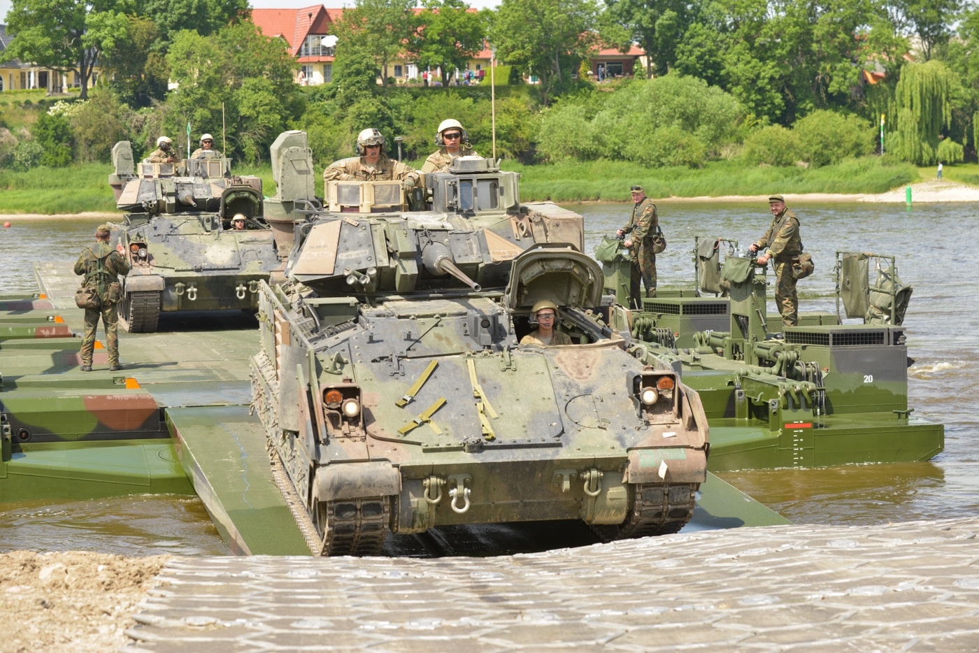 This image shows multiple M2A3 Bradley Infantry Fighting Vehicles from the 3rd U.S. Infantry Division crossing the Elbe River during the 2015 exercise Heidesturm Shock near Storkau, Germany. The Bradleys are positioned on a mobile bridge or ferry, moving over a wide section of the river with water visible on both sides. Soldiers are visible on or near the vehicles, observing the crossing and managing equipment. The background includes a view of the opposite riverbank with green foliage and clear skies, illustrating a typical European landscape. The vehicles are painted in standard camouflage and are equipped with upgraded armor and electronics distinctive to the M2A3 model. This exercise was designed to test and demonstrate the unit’s ability to conduct complex river crossings, a critical skill in NATO operations. The photo highlights the importance of mobility and adaptability for mechanized units in modern military training.