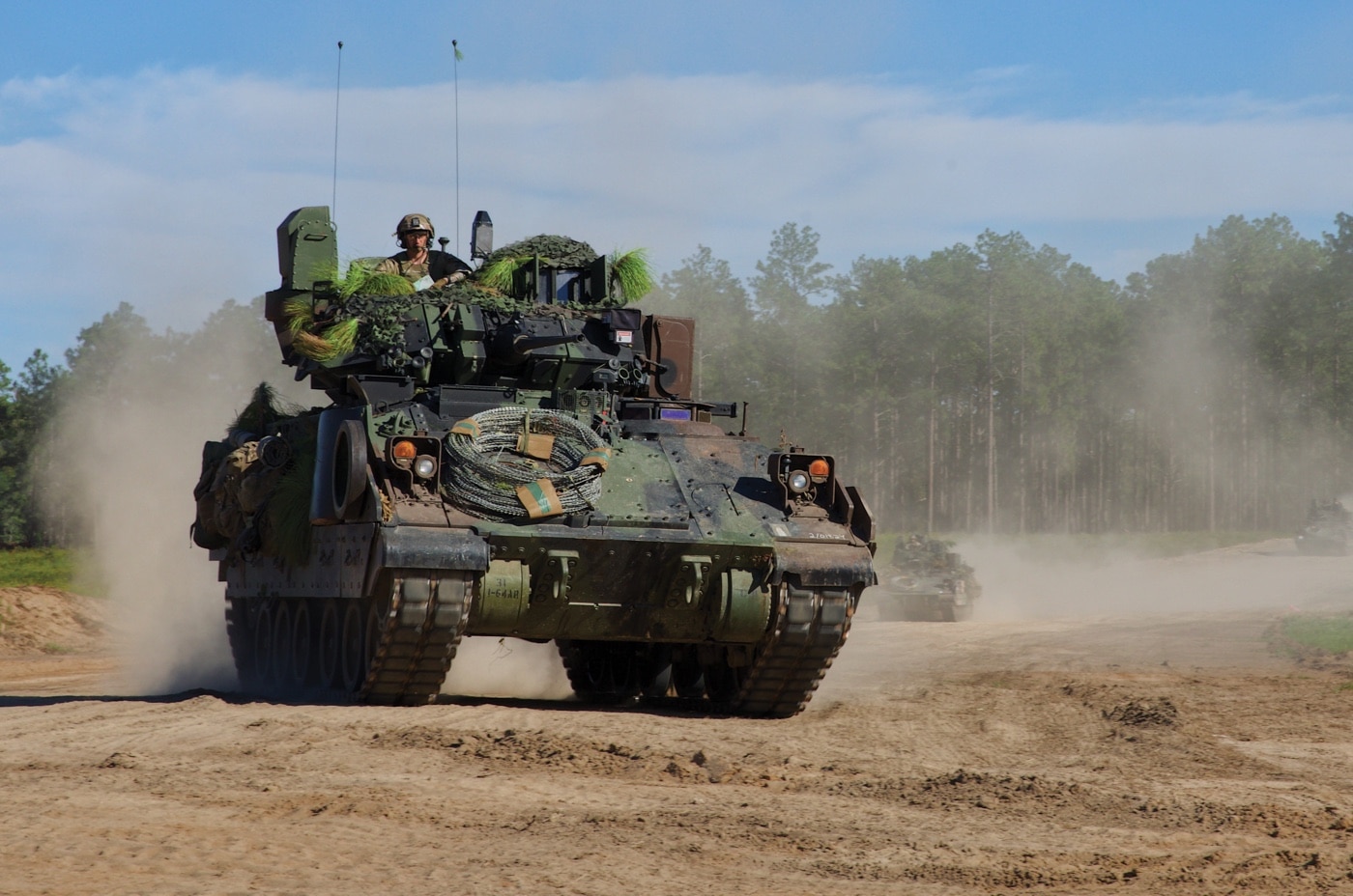 U.S. Army Soldiers with the 3rd Infantry Division are photographed participating in field training with M2A4 Bradley infantry fighting vehicles during Joint Readiness Training Center rotation 23-10. The image shows several Bradley vehicles in a training area, surrounded by soldiers in full combat gear preparing for a scenario. Some soldiers are on foot, coordinating movements, while others remain inside or near the vehicles. The terrain features grass, dirt, and scattered vegetation typical of JRTC at Fort Johnson, Louisiana. The M2A4 Bradley’s upgraded turret and additional armor are visible, reflecting recent modernization efforts. This training event simulates combat conditions, focusing on coordination between mechanized infantry and their vehicles. Scenes like this highlight the continuous evolution of tactics and technology in U.S. Army operations.