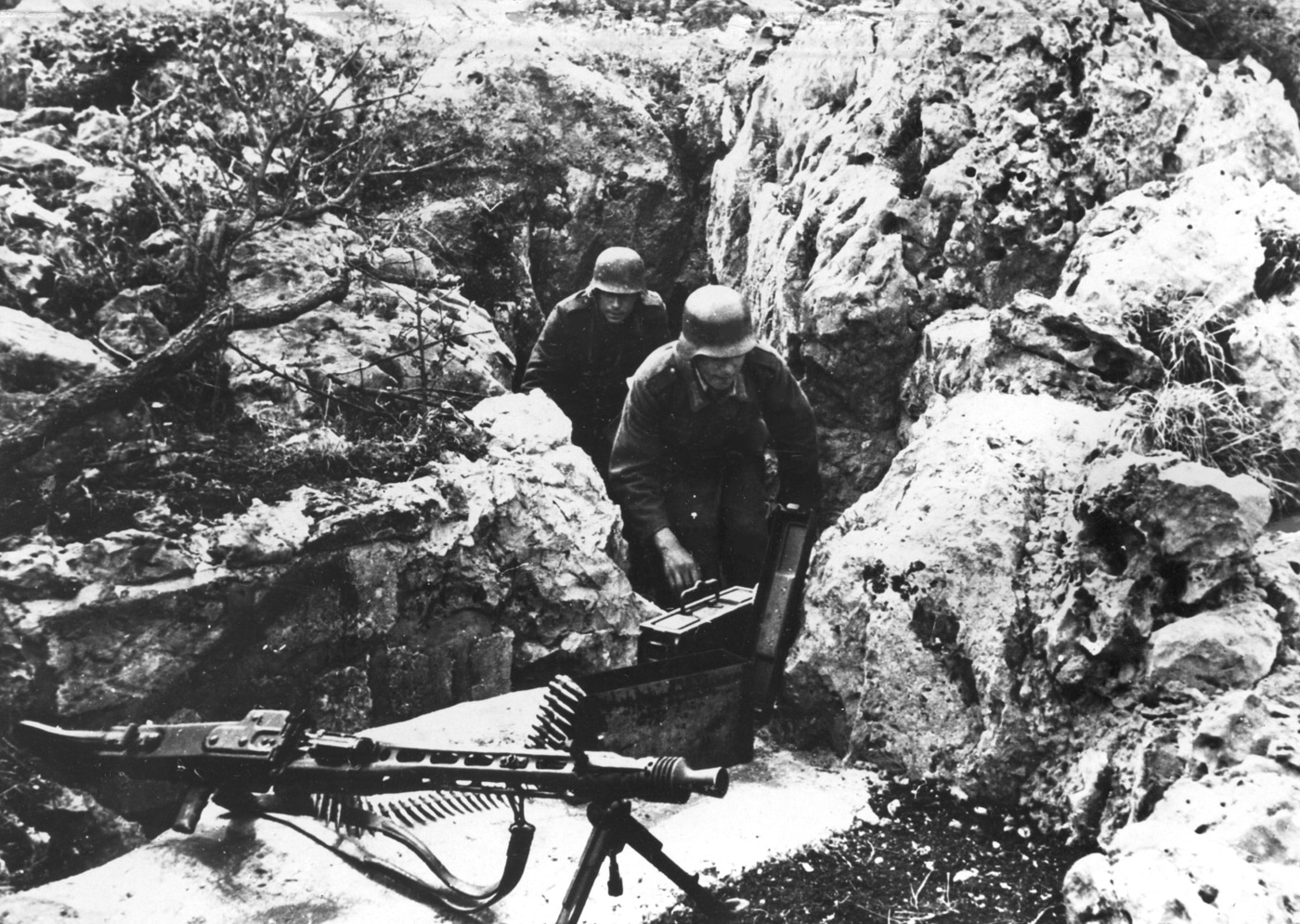 An MG 42 heavy machine gun is set up in a carefully prepared firing position nestled between large rocks on a mountain slope along the Italian front. The weapon is mounted on its bipod, aimed out over rugged terrain, with sandbags and stacked stones reinforcing the site. Nearby, there are supplies and ammunition boxes for quick access during combat. The scene shows how German troops used the natural landscape to protect the MG 42 crew and maximize the weapon’s field of fire. This image highlights the challenges and tactics of mountain warfare with the MG 42 machine gun.