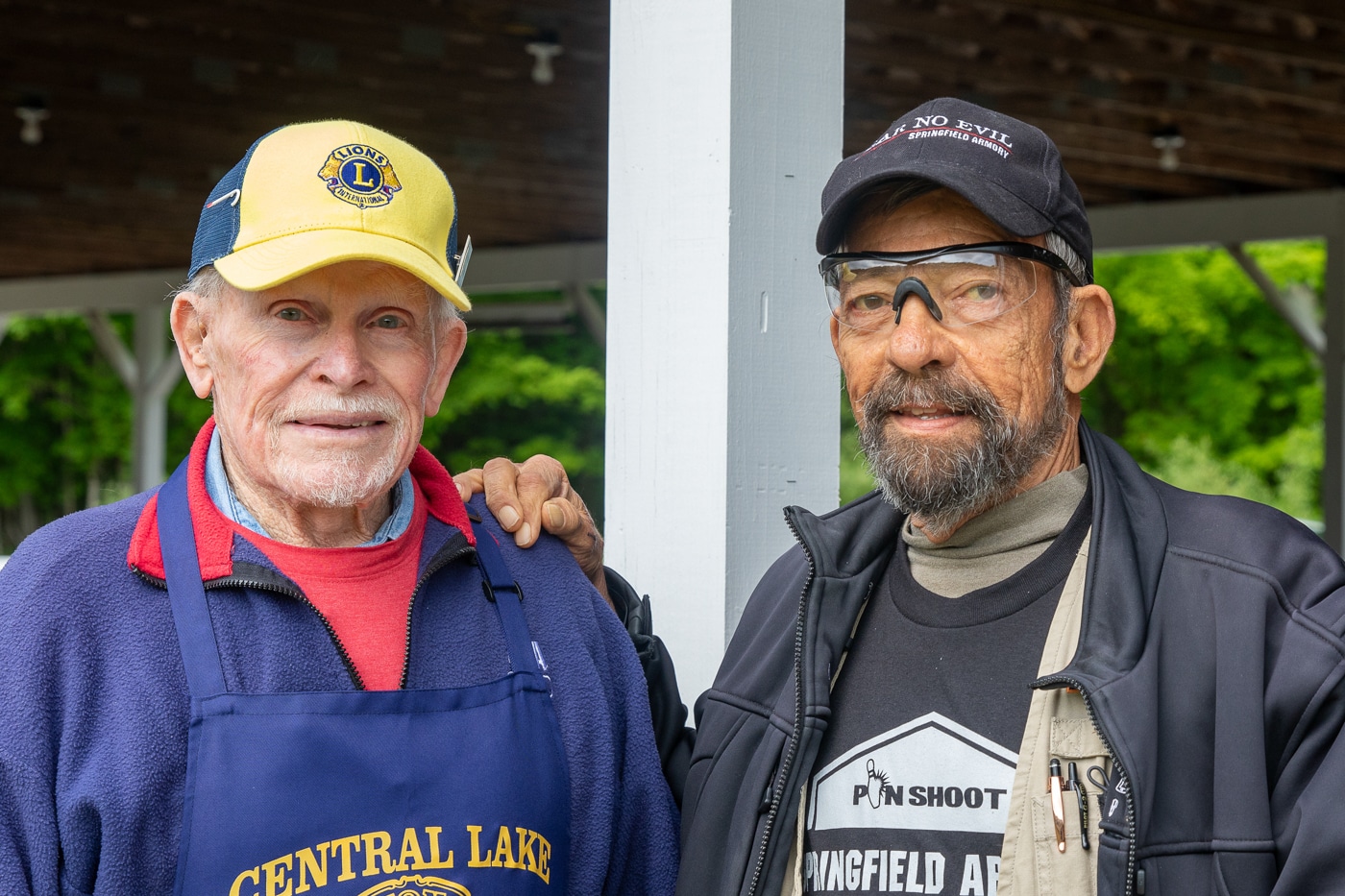 In this image, Al Burnett, recognized as a many-time national shooting champion, stands on the left next to author and photographer Massad Ayoob. The two men are posing together after the Springfield Armory Shootoff, smiling for the camera. Burnett is noticeably older, with a welcoming expression, while Ayoob stands beside him, dressed casually. The photo appears to be taken at the event venue, possibly outdoors or in a well-lit indoor area. Both men are wearing casual or event-related attire, highlighting their connection to the match. The atmosphere is relaxed and friendly, reflecting the camaraderie of the shooting sports community. This image documents two notable figures at the 2025 Springfield Armory Shootoff.