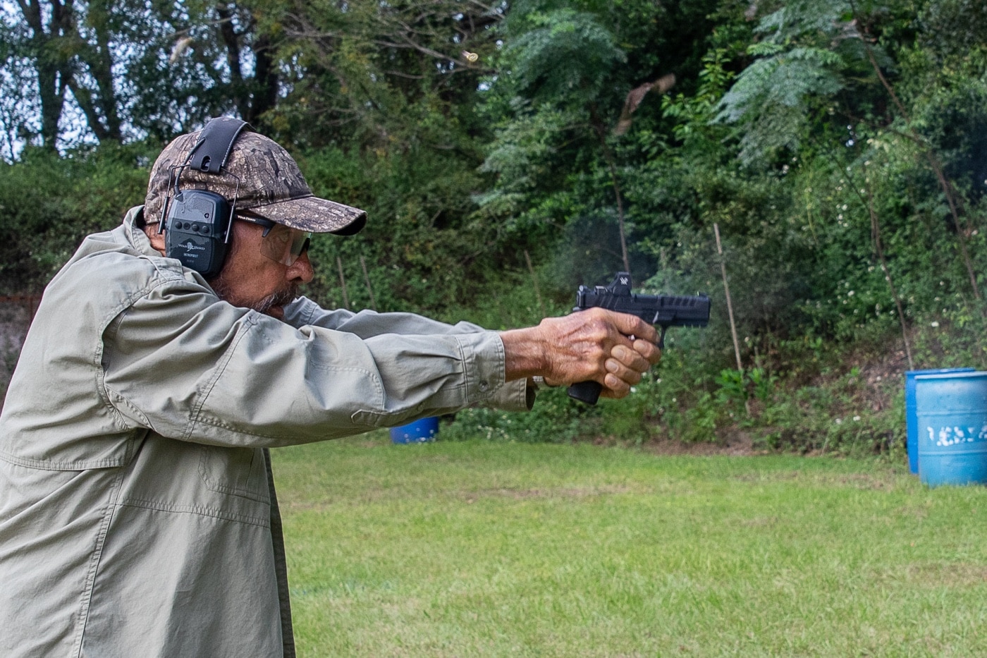 The photo shows Massad Ayoob in the act of demonstrating controlled trigger slap with an Echelon 9mm semi-automatic pistol. He is gripping the pistol firmly, with his trigger finger quickly coming off the trigger after firing, illustrating the speed-focused technique described in his article. The image captures the dynamic moment of rapid fire, showing Ayoob’s form, stance, and the visible motion of his finger. Range safety gear such as ear and eye protection is worn, and the background includes a standard shooting range setup. This visual supports Ayoob’s point about the usefulness of controlled trigger slap for close-range, high-speed shooting, especially when accuracy is not as critical as fast follow-up shots. The photo highlights the aggressive nature of this trigger control method. It also reinforces the importance of proper grip when using techniques that sacrifice finesse for speed.