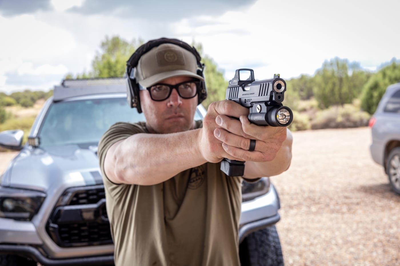 A person is pictured on an outdoor shooting range, standing in a proper stance and aiming a semi-automatic pistol at a paper target downrange. The shooter’s finger placement and grip show attention to technique, reflecting instruction on trigger control. The scene includes typical range safety features, like safety glasses and earmuffs. This image illustrates live-fire practice, where shooters work on manipulating the trigger smoothly to improve accuracy. The context ties directly to Massad Ayoob's teaching on different trigger manipulation methods, including trigger weld and riding the link. The shooter is mid-practice, showing focus and concentration on each shot. The environment is clear and uncluttered, emphasizing the technical aspects of shooting technique.