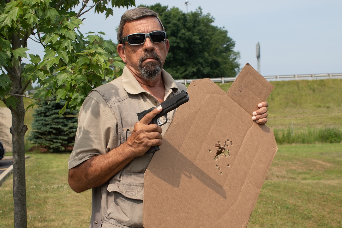 The photo shows Massad Ayoob standing next to a large paper shooting target, pointing out specific areas that were hit during a firearms training class. He is in an outdoor or indoor range setting, surrounded by training materials and wearing typical range gear such as ear and eye protection. Ayoob’s posture suggests he is explaining shot placement, accuracy, and the importance of controlled fire to a group of students. The target displays various bullet holes, illustrating outcomes of live-fire drills often used to teach self-defense marksmanship. This image connects to topics about firearms training, defensive shooting skills, and the documentation of ability for legal defense in self-defense cases.