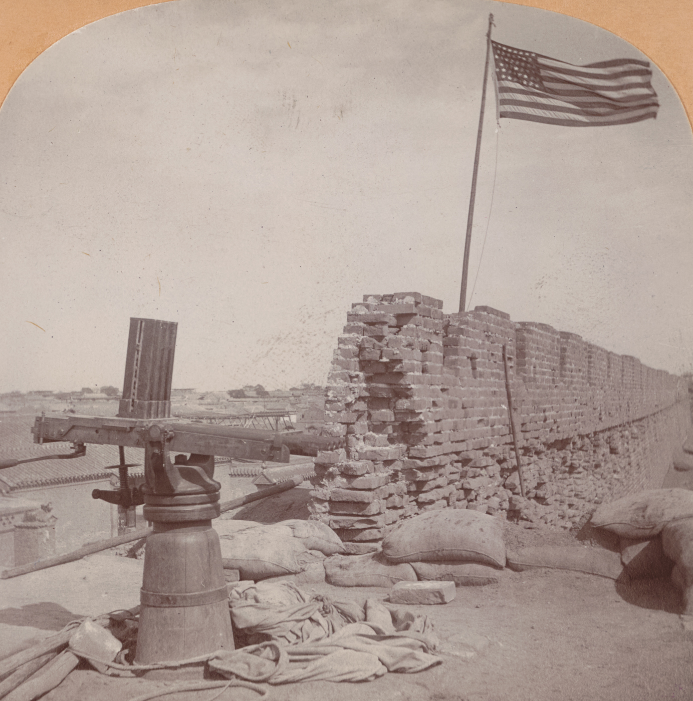 The photograph shows a Nordenfelt gun, a multi-barreled rapid-fire weapon from the late 19th century, positioned on the city wall near the Great South Gate of Tien-Tsin (Tianjin), China. The gun, mounted on a simple wheeled carriage or tripod, points out over the city or across the defensive ramparts. Surrounding the weapon are sandbags, debris from battle, and the thick stone parapet of the wall itself. Nearby, members of the 9th U.S. Infantry, in field uniforms and campaign hats, stand guard or examine their newly captured prize. The scene reflects the aftermath of a successful assault by American forces during the Boxer Rebellion. The background may show distant city buildings, smoke from recent fighting, or additional fortifications. This image highlights the use and capture of modern weapons in urban combat and the strategic importance of city gates and walls in the Tien-Tsin campaign.