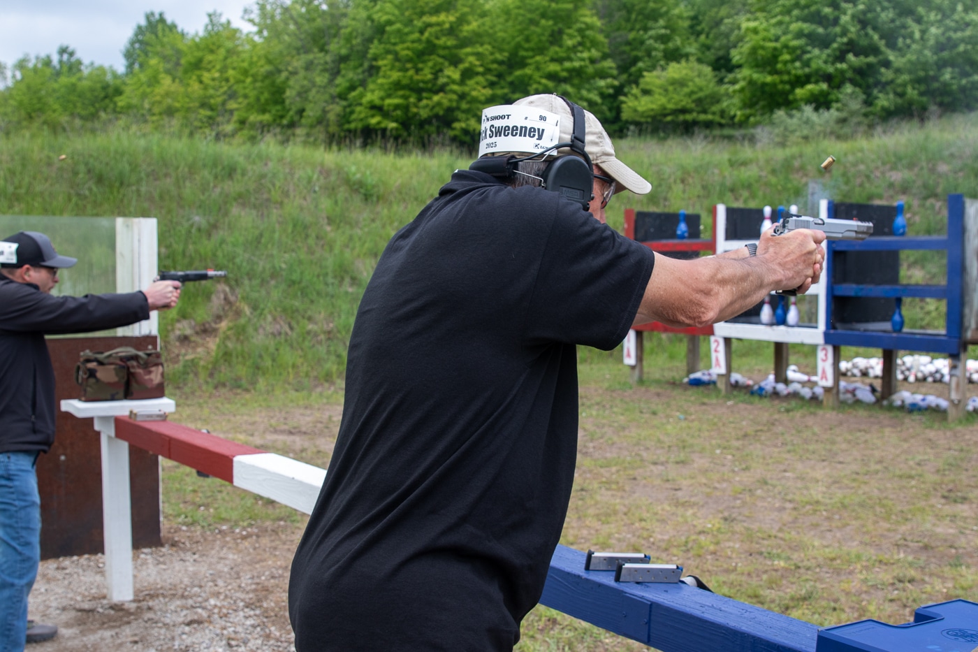 The photo shows Patrick Sweeney in mid-action at the Springfield Armory Shootoff, aiming and firing his Springfield Armory .45 caliber pistol at a set of bowling pin targets. Sweeney is in a solid shooting stance, demonstrating focus and control as he competes for a place in the finals. The image captures the moment of recoil, with a spent casing possibly visible in the air and smoke near the muzzle. The background includes parts of the outdoor range, such as shooting tables, pin targets, and other competition equipment. Sweeney is wearing appropriate safety gear, like shooting glasses and hearing protection. The scene is brightly lit, highlighting the intensity of the match. This action shot illustrates Sweeney’s skill and determination as he advances toward the championship round.