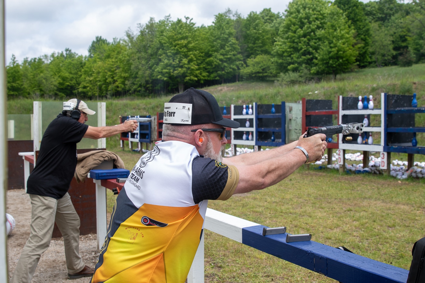 The photo captures the final moments of the Master Blaster shootoff at the Springfield Armory event. Phil Farr is in the foreground, standing in a shooting stance and aiming a .50 GI pistol at the target downrange. Behind or to the side, Pat Sweeney is visible holding and firing his .45 caliber handgun, also aiming at his targets. Both shooters are intensely focused, with their attention fixed on the competition. The background shows elements typical of a shooting range, including target tables and possibly a few spectators or range officials. The atmosphere is tense, marking the climax of the competition. Farr and Sweeney are dressed in appropriate match attire, with safety gear visible. The image documents the high-stakes final duel that decided the Master Blaster champion at the 2025 Springfield Armory Shootoff.